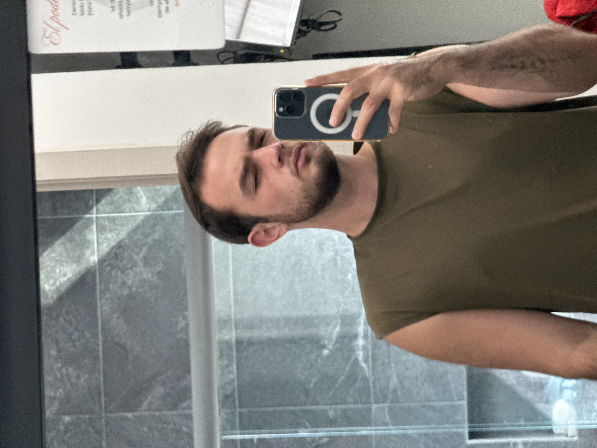 A man is taking a selfie in a bathroom. The bathroom has gray tiled walls and a glass shower door. The man is wearing a green t-shirt. He is holding an iPhone with a black case and a visible MagSafe ring. The background includes a partially visible document with Spanish text. The image was likely taken indoors, possibly in San Bernardino Tlaxcalancingo, Mexico.