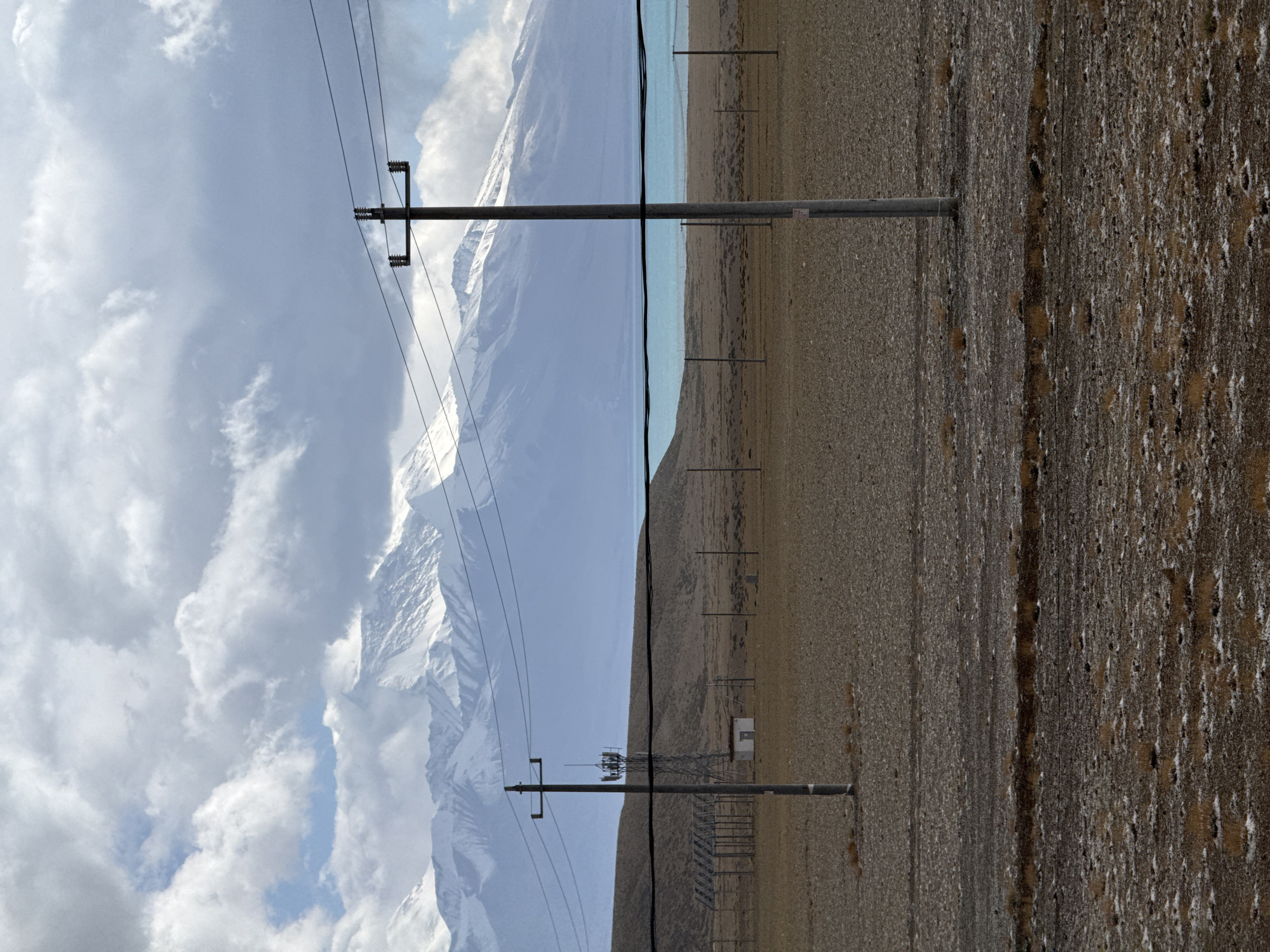 The image depicts a landscape in Hor town, China, featuring a snowy mountain range on the left under a cloudy sky. A body of water separates the mountains from a barren, brown landscape dotted with electrical poles and wires, stretching towards the right of the frame. Some poles are of a taller, lattice-style construction. The ground is dry, and its coloration indicates a sparse vegetation cover. The image is taken from a vantage point that positions the electrical poles as prominent foreground elements against the backdrop of the mountain range.