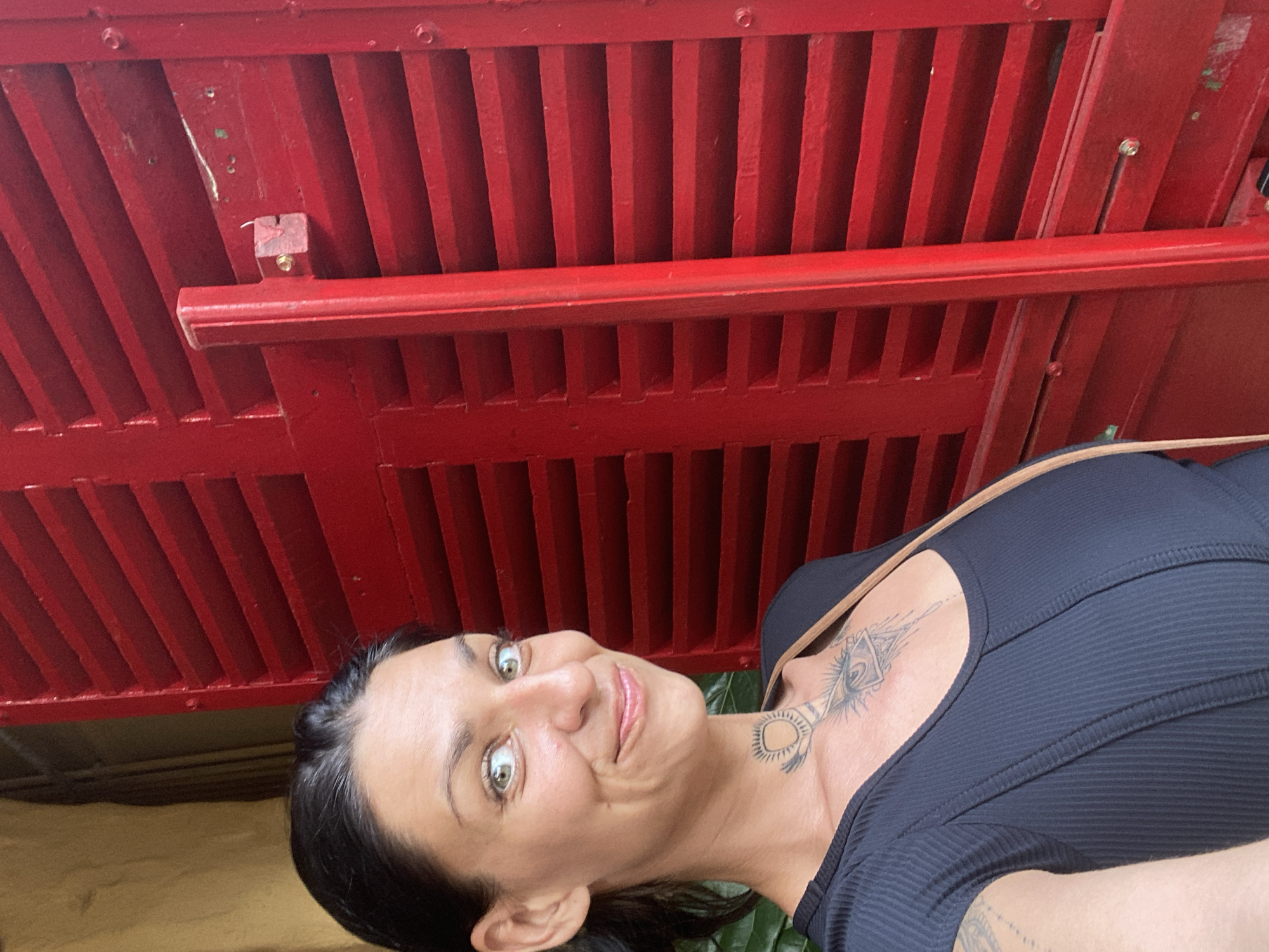 A woman with dark hair and green eyes is standing in front of a red shuttered wall. She is wearing a black shirt and has tattoos on her neck and chest. She is smiling and looking at the camera. The environment appears to be outdoors, possibly in the city of Mũi Né, Vietnam, given the style of the shutters.