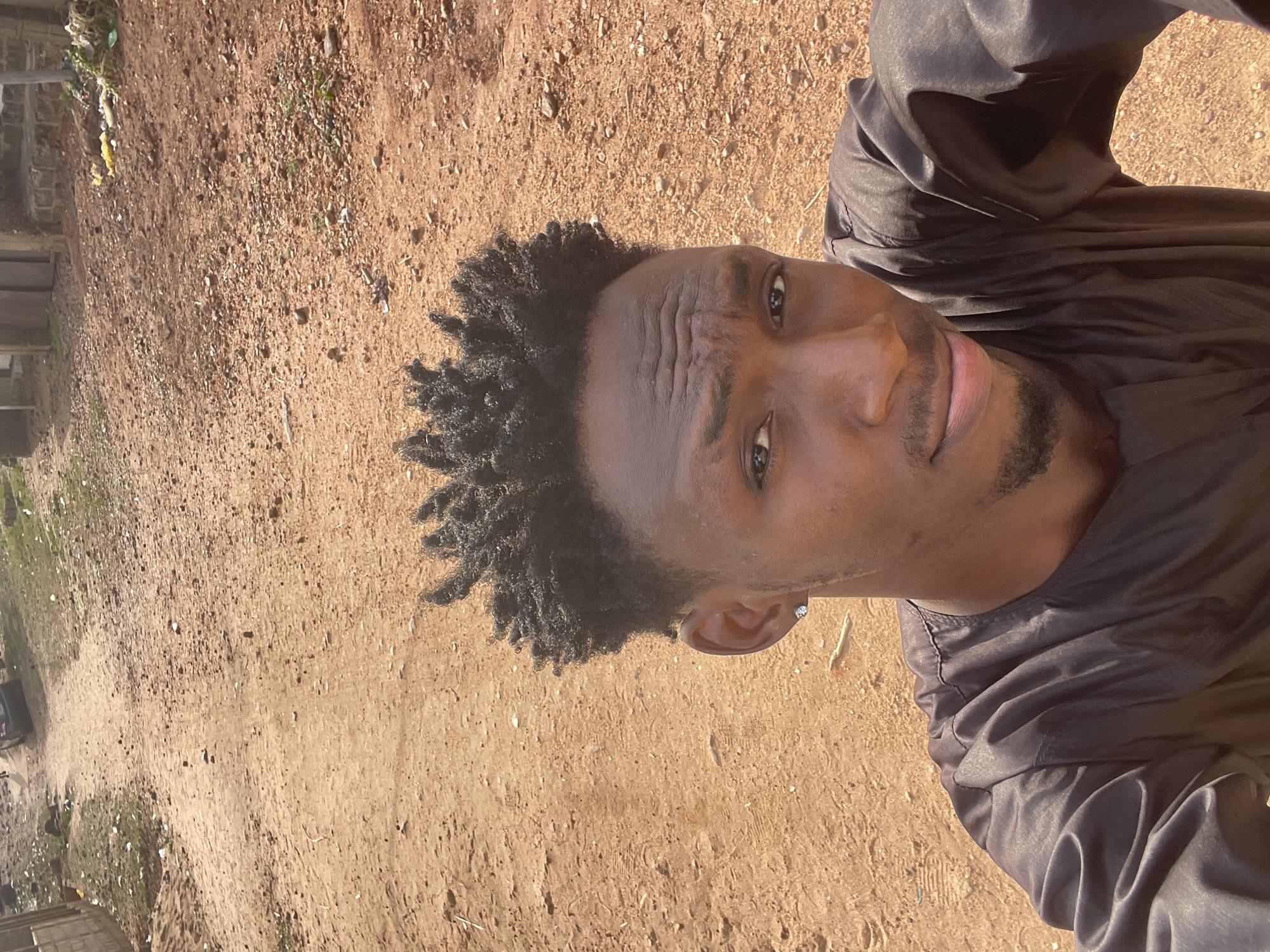 The image shows a man in an outdoor setting in Lokoja, Nigeria. He is wearing a black top and has short, textured black hair. The ground is dirt with small rocks and sparse vegetation visible in the background. The background also includes parts of wooden structures and some refuse.