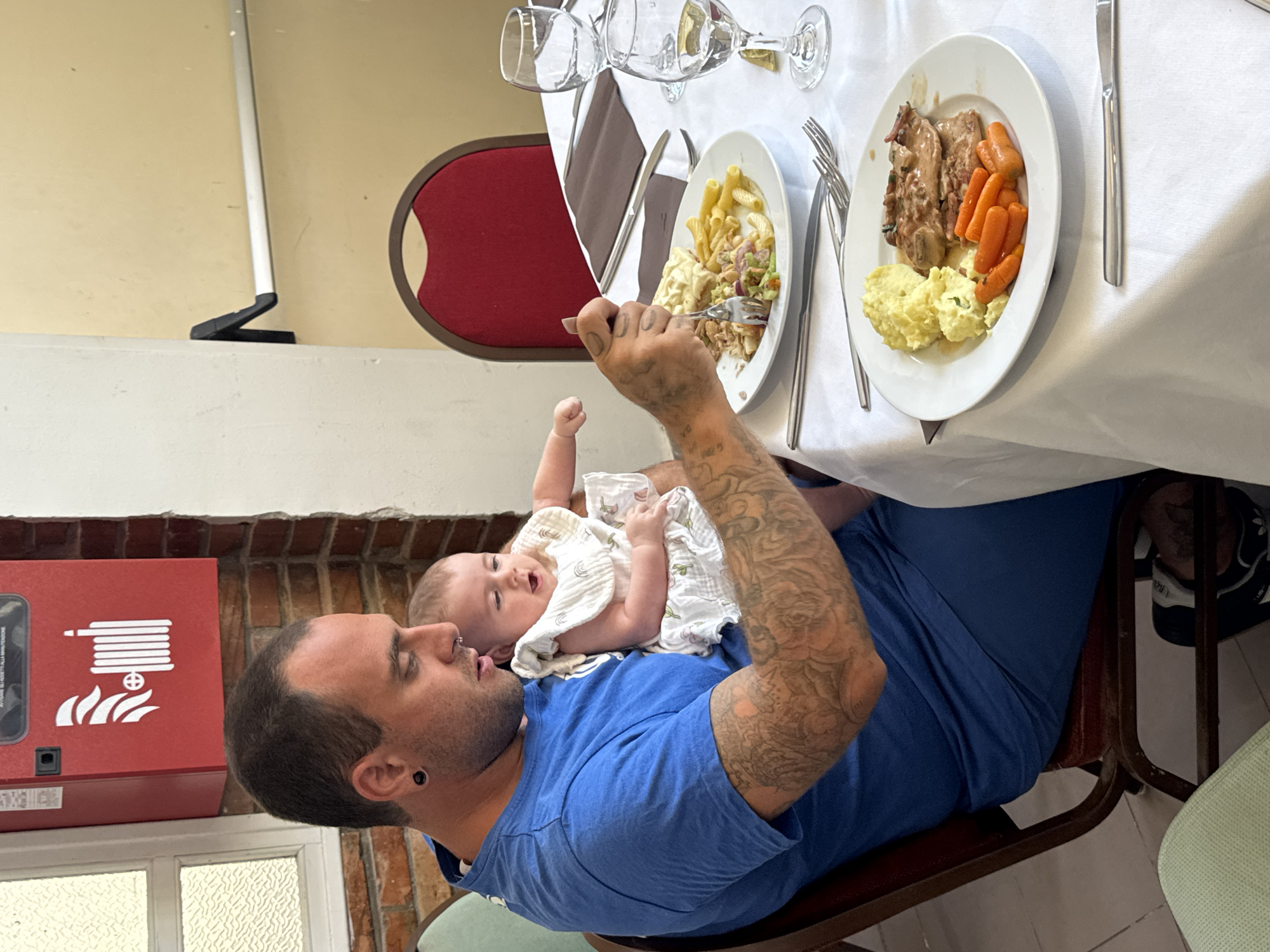A man in a blue t-shirt holds a baby wearing a white dress while sitting at a table. The man has tattoos on his arms and is using a fork to eat from a plate of food. The baby appears to be looking at the man. The table is covered with a white tablecloth and set with plates, glasses, and cutlery. In the background, there is a red fire extinguisher mounted on a brick wall, and a red chair with a curved back. The overall setting suggests a restaurant or dining hall in Capaci, Italy.