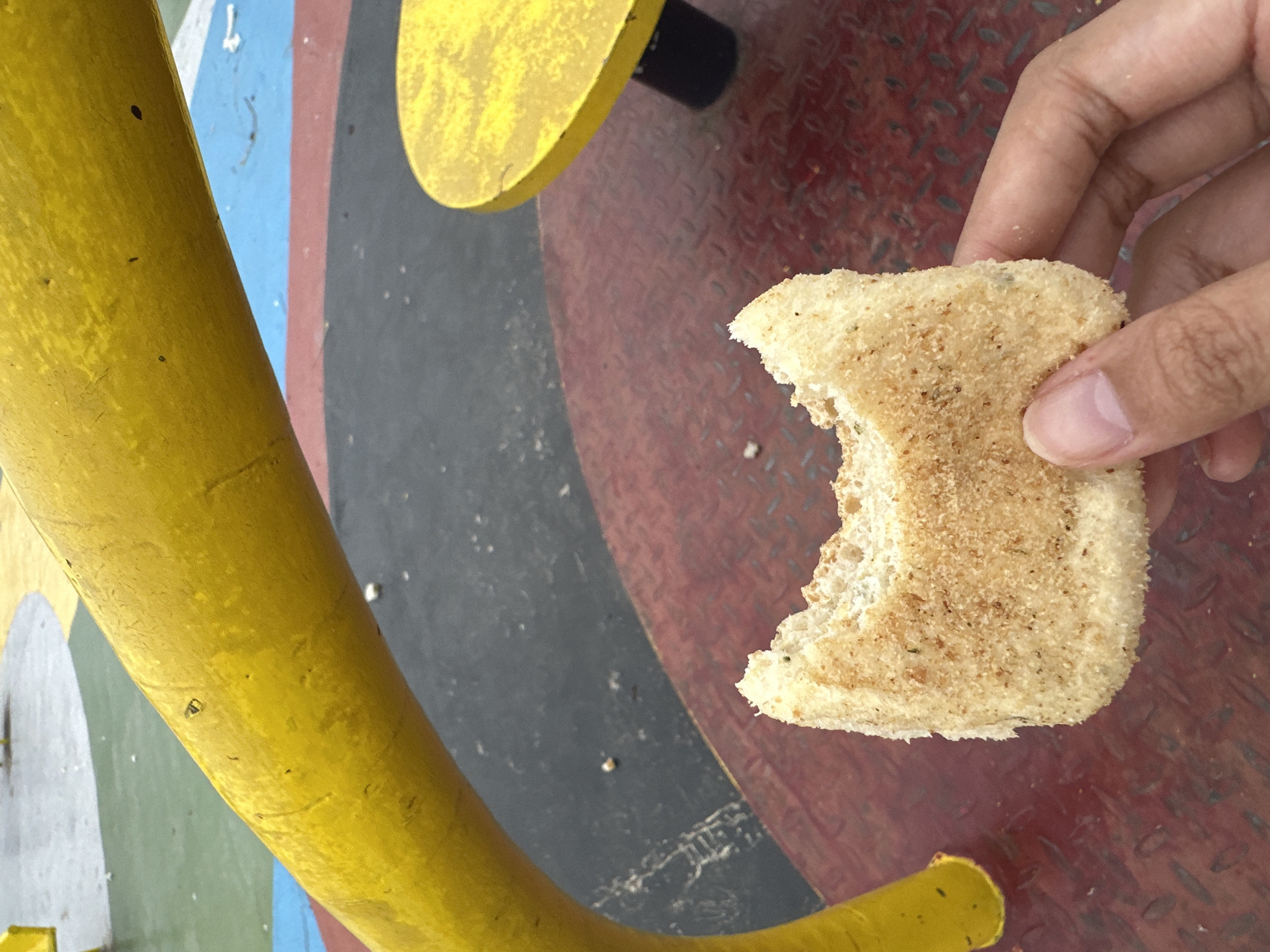 An overhead shot captures a human hand holding a piece of light brown, seasoned bread with a bite taken out of it. The bread appears toasted or fried, covered in fine crumbs and dark specks, possibly herbs or spices. The hand's thumb and fingers are visible, with neatly trimmed nails.
The setting is an outdoor, brightly lit public space, likely a park or recreational area in Davao City, Philippines. The background features a vibrant, multi-colored ground surface, painted with geometric or abstract shapes in red, black, blue, yellow, and white/gray. A prominent circular section in red displays a textured, diamond-plate pattern, suggesting a durable metal surface.
To the left, a bright yellow, thick, curved metal structure, possibly a pipe, railing, or part of playground equipment, runs vertically along the edge of the frame. Another smaller, flat yellow circular element is visible in the upper center, connected to a black cylindrical piece. The overall scene suggests a casual moment of eating in an urban environment with colorful, functional design elements. No text is visible in the image.
