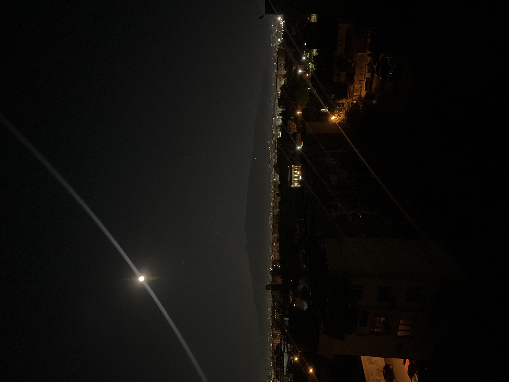 The photograph, taken at night, shows a cityscape under a dark sky. The moon is visible, with a light streak crossing the sky. Along the horizon line, there are city lights from buildings and streets. The foreground contains dark building structures and power lines. Given the city lights and general layout, the location is possibly Sofia, Bulgaria. No people are visible.