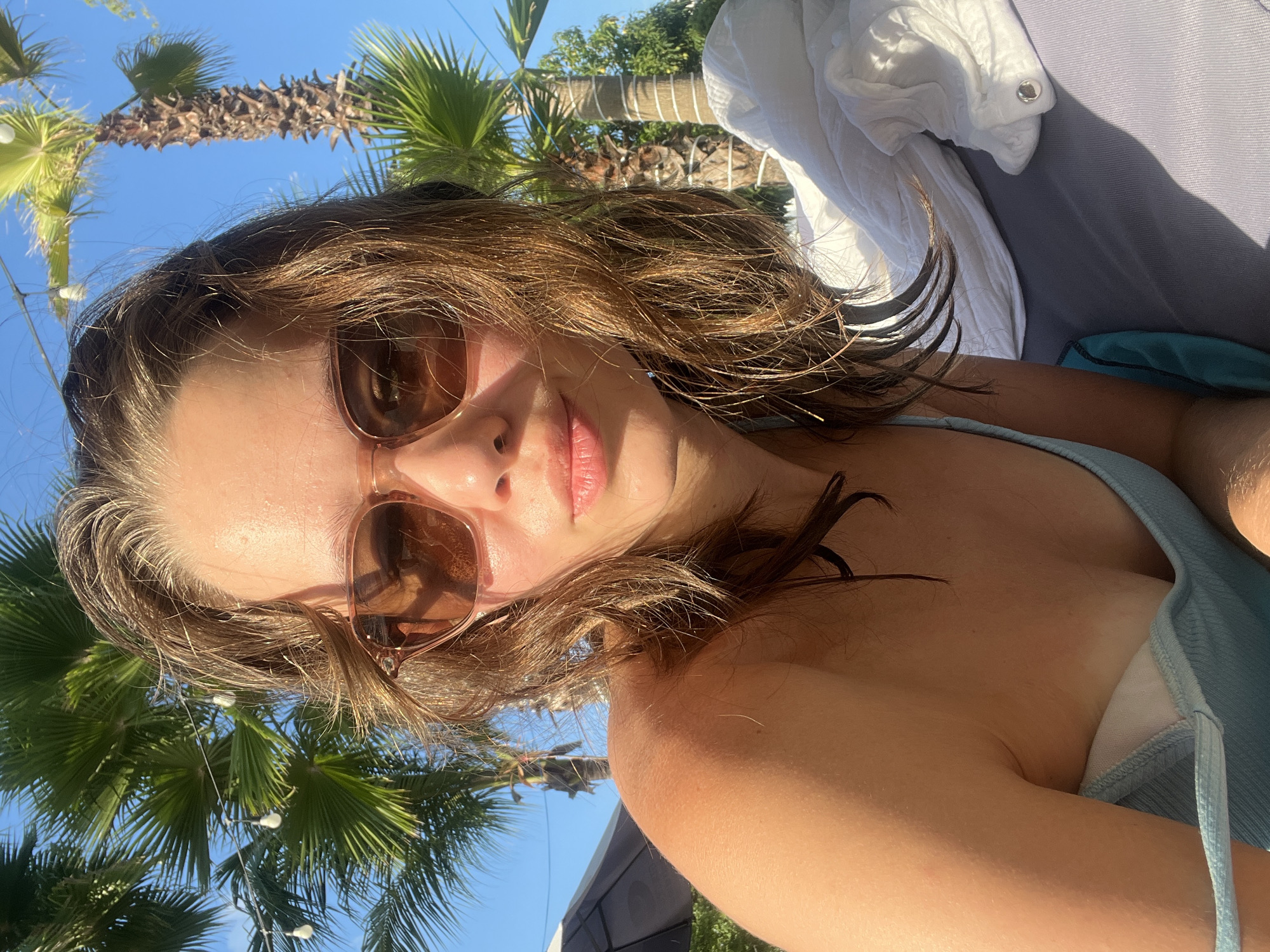 A woman is wearing sunglasses outdoors. Palm trees and a blue sky are visible in the background. The woman appears to be in a tropical location. The location could potentially be in Nam Nha Trang Ward, Vietnam.