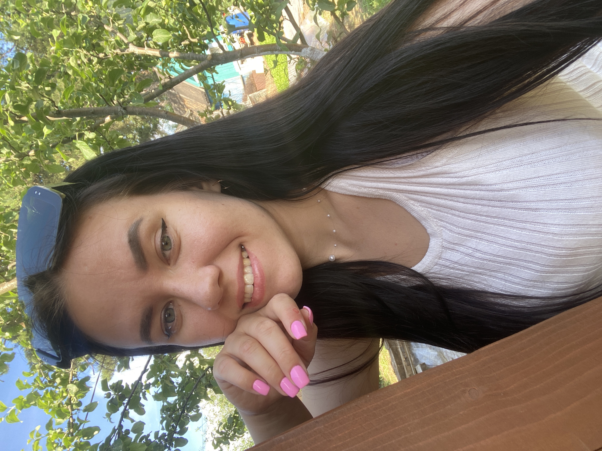 An outdoor portrait, set in the city of Chaikovsky, Russia, depicts a young woman smiling directly at the viewer. She possesses long, dark, straight hair, light-colored eyes enhanced with black winged eyeliner, and a silver nose piercing on her left nostril. Dark sunglasses are positioned on top of her head. Her nails are painted a vibrant pink. She wears a light-colored, possibly white or pale pink, ribbed tank top and a delicate necklace with small pearl-like beads. Her left hand, with fingers curled and nails visible, supports her chin, resting on a prominent brown wooden railing in the lower-left foreground. The background features lush green foliage from trees under a bright blue sky. Portions of a distant structure, characterized by a teal-colored wall and a red roof, are discernible through the trees. The scene is illuminated by natural daylight.