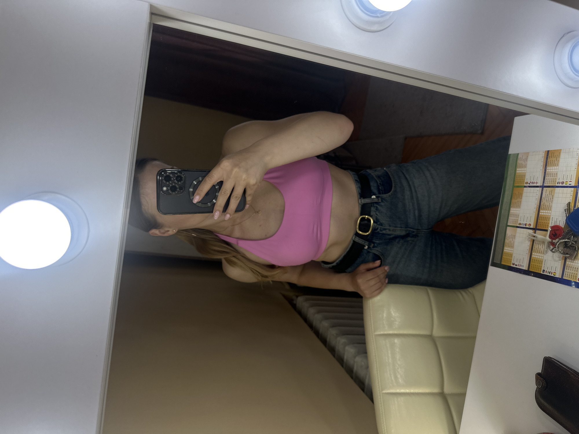 A person is captured in a mirror selfie, seen reflected in a brightly lit vanity mirror. The person, a woman with long blonde hair, is wearing a vibrant pink crop top, blue denim jeans, and a black belt with a prominent gold buckle. She holds a dark-colored smartphone, likely an iPhone given the distinct camera bump and MagSafe-like ring, in her left hand, obscuring most of her face. Her nails are manicured with a light pink polish, and a delicate gold necklace is visible around her neck.
The person appears to be sitting on a light-colored (cream or off-white) tufted chair. The mirror itself is framed with multiple circular lights, illuminating the scene. To the right of the reflected person, on a table or shelf, are several objects: a sheet of paper with a grid and text, a set of keys with a red keychain, and a dark brown wallet or small purse. The text on the paper includes abbreviations for days of the week in Ukrainian ("ПН ВТ СР ЧТ ПТ СБ НД"), along with numbers such as "+020" and "020+", suggesting it might be a schedule or calendar. The background, visible in the mirror's reflection, shows an indoor setting with a dark wall or door and a radiator. The activity is a casual mirror selfie, likely taken in a personal space such as a dressing room or bedroom.
