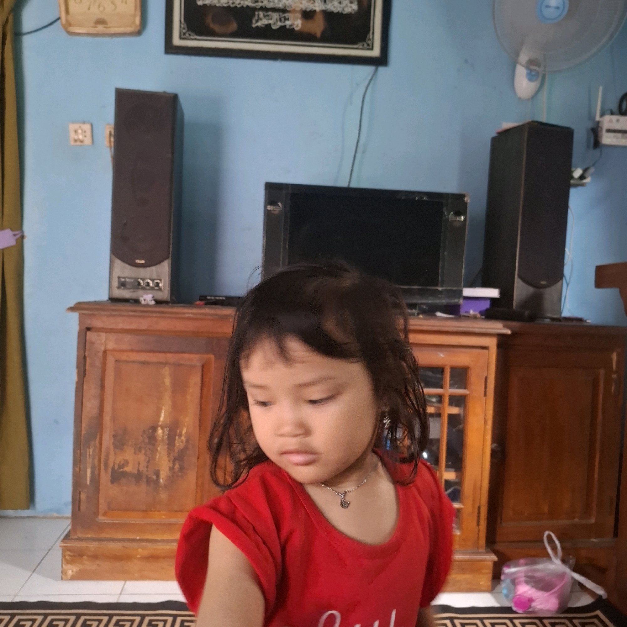 A young girl in a red t-shirt stands in front of a wooden cabinet. She is looking down and to her right. Behind her is a television set and two speakers. To the left of the cabinet, a curtain hangs. Above the cabinet on the blue wall, a clock and a framed picture are visible. A ceiling fan is mounted on the wall to the right. The room appears to be a living room.