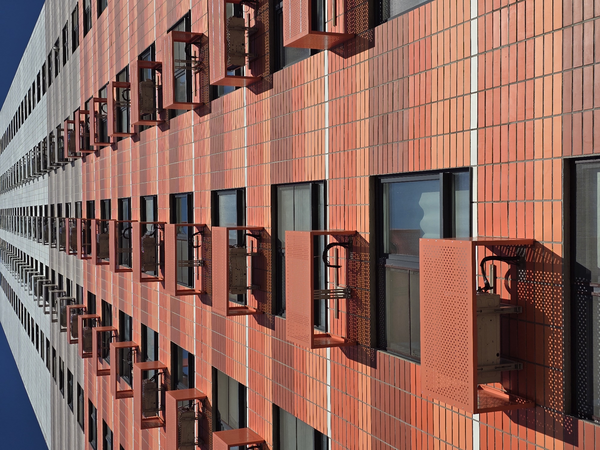 The image depicts a modern building in Moscow, Russia, characterized by a two-toned facade. One section of the building is clad in orange and red vertical tiles, while the other is gray and white. Windows are spaced regularly across both sections of the building. Attached to the windows on the orange-red facade are perforated, reddish-brown metal boxes or enclosures. These structures appear to house air conditioning units. The image is taken from a low angle, emphasizing the height of the building. The weather appears to be sunny and cloudless.