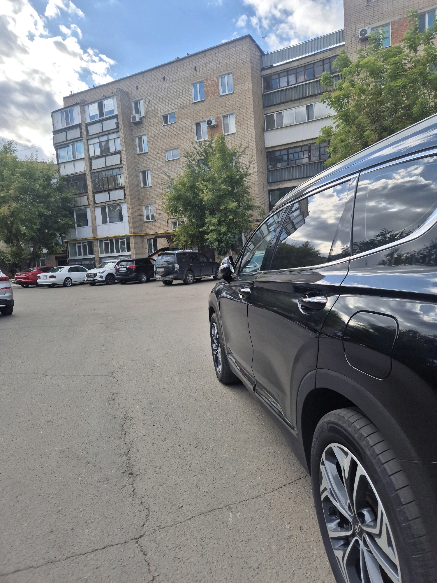 A black SUV is parked in a lot in front of a multi-story brick apartment building under a cloudy sky. The building has several balconies and windows. Several other cars are parked in the lot, including a red sedan, a white sedan, and other SUVs. Trees are visible around the building. The ground is paved and cracked.