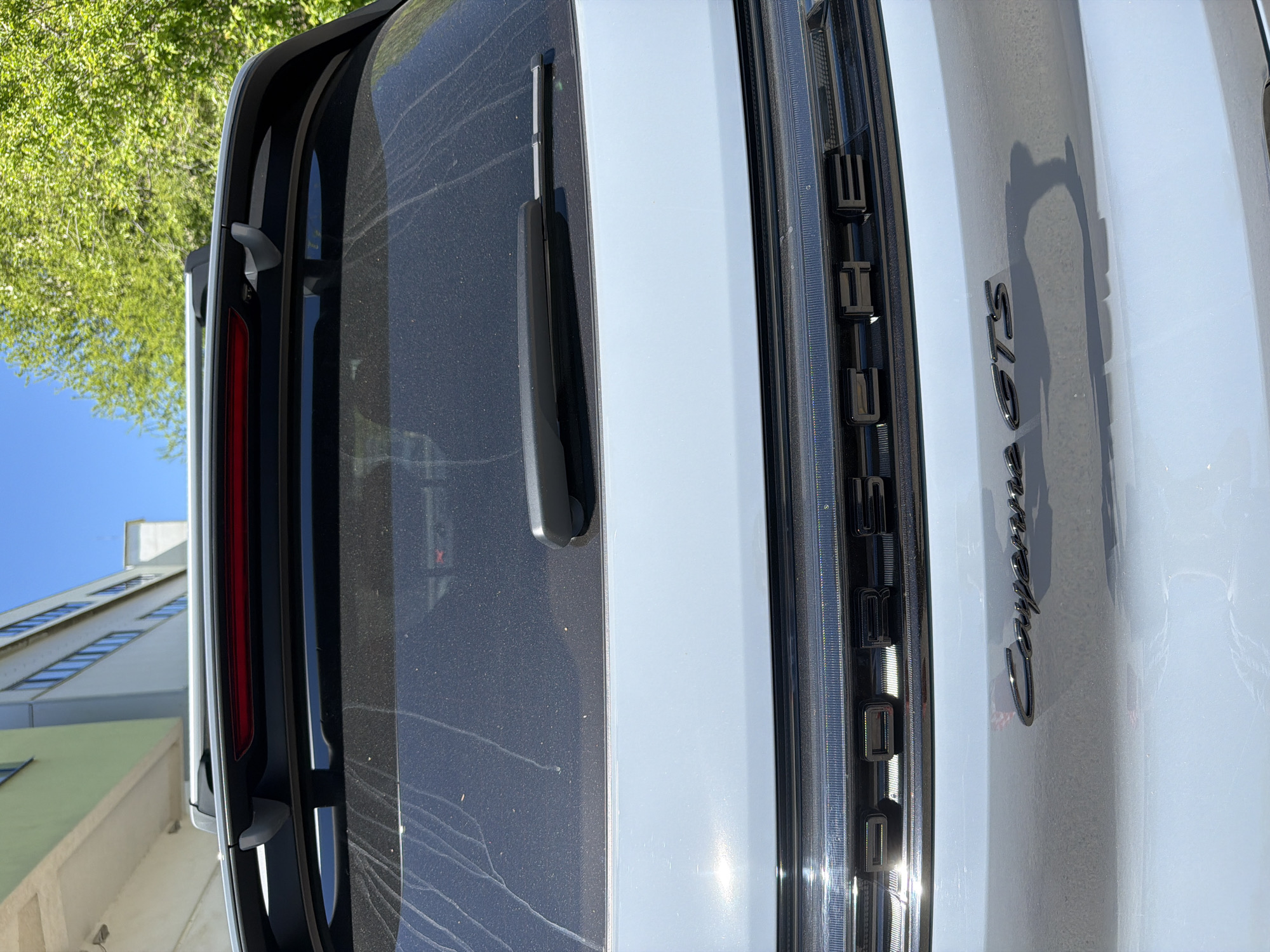 The image depicts the rear of a white Porsche Cayenne GTS. The words "PORSCHE" and "Cayenne GTS" are visible on the vehicle's back. The scene includes a rear windshield wiper and a portion of the tail light. In the background, there are trees, the sky, and a multi-story building. The precise location is not discernible from the image, although Bulgaria is listed as a possible location.