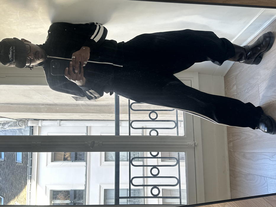 A male individual is captured in a full-length mirror selfie, positioned indoors. He has a dark complexion, wears dark-framed glasses, and sports a moustache and goatee. His head is covered by a dark cap featuring a light-colored car illustration and partial text "L'ART DE L'AUTOMOBILE EST DE...". He is dressed in a black tracksuit; the jacket has white stripes on the sleeves and is partially unzipped, and the loose-fitting pants display a single white stripe down the outer seam of the left leg. The individual wears a dark leather shoe on his left foot and a dark leather boot with a buckle on his right foot. Rings are visible on both hands. He holds a dark smartphone in both hands, directing its camera towards the mirror. The indoor setting features light-colored wooden floorboards and a plain white wall. Behind the subject, a tall, white-framed casement window looks out onto classic Parisian architecture. Visible outside are light-colored Haussmannian buildings with multiple windows, ornate black wrought-iron balconies, and mansard roofs, all characteristic of a city location in Paris, France. A decorative black wrought-iron balcony railing is also visible through the lower section of the window.