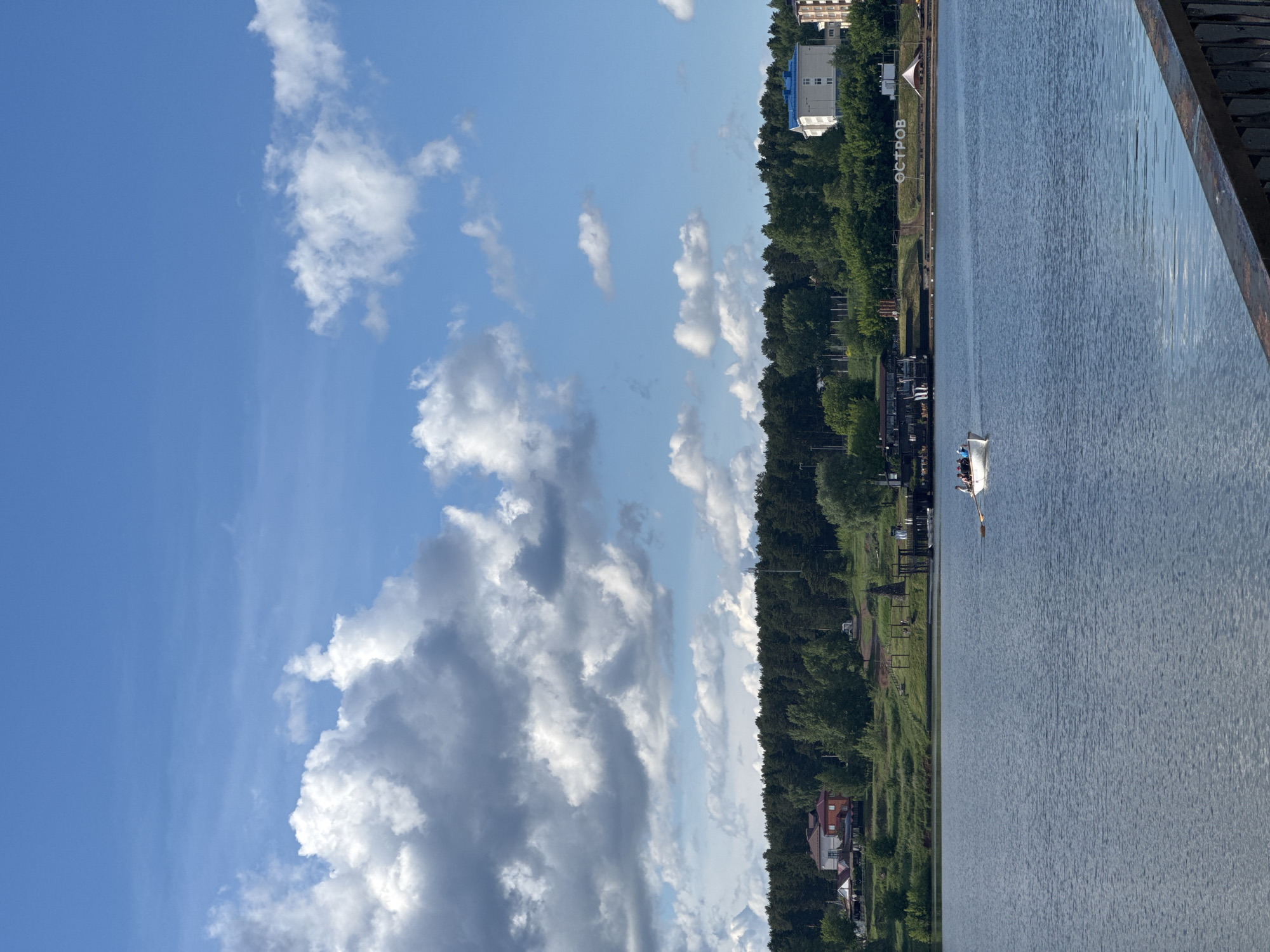 The image depicts a sunny day at a river or lake in what appears to be a developed area, possibly in Votkinsk, Russia. The view is oriented sideways. A body of water dominates the right side of the image, with the reflections of the sky and some landmass visible. In the water, there is a white rowboat with two visible people rowing. A tree line runs along the edge of the water on the left, with buildings intermittently visible along the shore. One notable structure is a large building with a blue roof and a few white buildings. A grassy area with a sign is along the shoreline. The sky occupies much of the left side of the image, filled with white cumulus clouds against a blue backdrop. Part of a metal structure is present in the top right of the frame.
