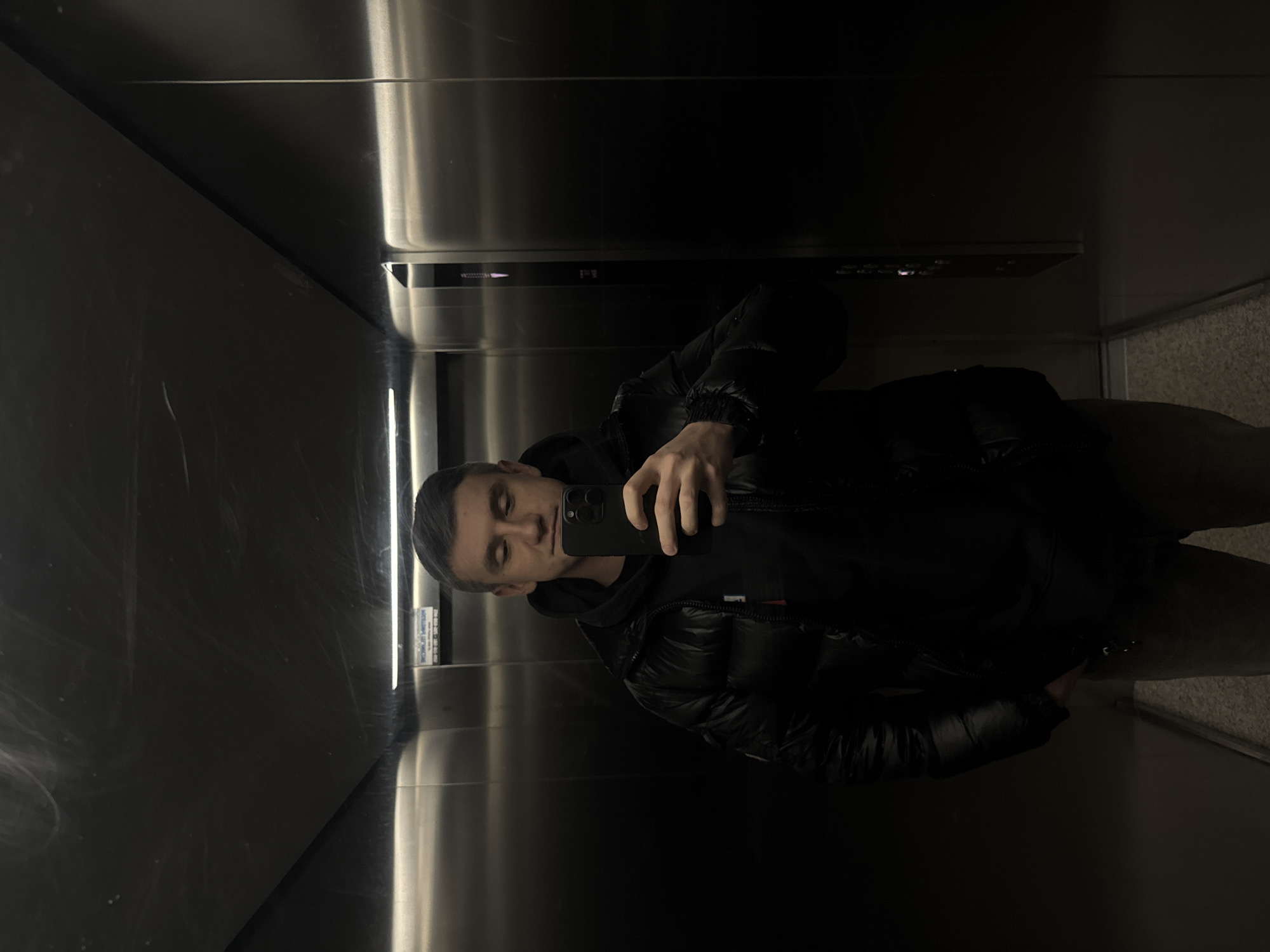A man stands in an elevator, holding a phone in his right hand to take a selfie. He is wearing a black hooded sweatshirt under a black puffer jacket. The elevator has stainless steel walls and a gray floor. Based on the visible sticker on the elevator wall, the location is in Warsaw, Poland.