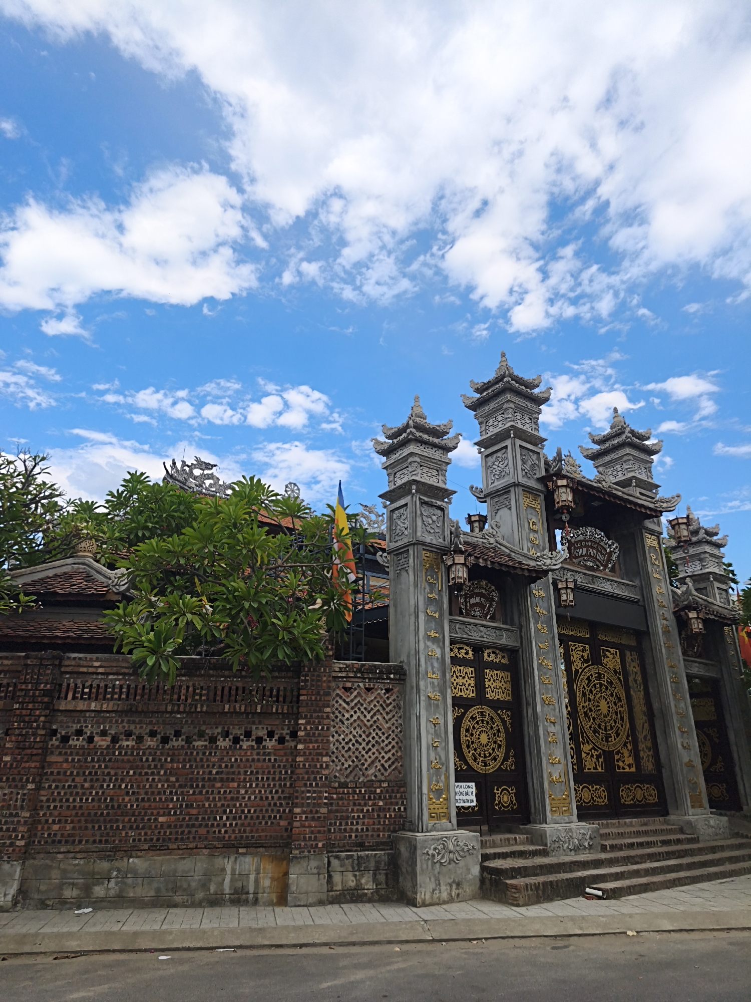The image depicts the ornate entrance to a temple or pagoda, likely in Nha Trang, Vietnam. The architecture features multiple tiered roofs with decorative finials, intricate carvings, and a grand set of imposing, dark double doors adorned with golden circular motifs. Flanking the entrance are tall, grey stone pillars embellished with sculpted details and smaller pagoda-like structures at their apexes. Hanging lanterns add to the atmospheric detail of the entrance.
To the left of the main gateway, a brick wall of varying textures and patterns stretches across the frame, topped with a lush green tree. Behind the wall, parts of the temple roof are visible, along with a dragon sculpture and a multi-colored flag. The overall impression is one of rich cultural heritage and spiritual significance.
The scene is set against a bright blue sky filled with scattered white clouds, suggesting it is daytime, likely mid-morning or afternoon, with pleasant weather. The ground in front of the entrance consists of stone paving and a road, indicating an urban or semi-urban setting. There are no people visible in the frame, focusing the attention solely on the architecture and the surrounding environment.
Visible text on a white sign hanging on the left door reads: "VUI LÒNG KHÔNG ĐI XE VÀO TRONG CHÙA". This translates to "Please do not ride vehicles inside the pagoda." Another circular sign above the entrance also appears to have text, but it is not clearly legible. The overall mood evoked is one of solemnity and visual splendor.
