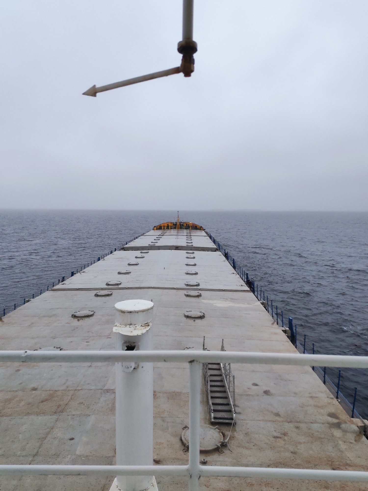 The image presents an overcast scene from the deck of a ship, likely a bulk carrier, at sea. The ship's deck stretches out towards the horizon, flanked by the dark expanse of the water. A railing runs along the edge of the deck, providing a sense of enclosure. A metal structure with an arrow-shaped indicator is visible overhead, possibly a part of the ship's navigation system. The deck is made of concrete and has a series of metal hatch covers running along its length. A set of stairs leads to a lower level on the deck. The sky is uniformly gray, suggesting a cloudy day at sea. The location in the city of Shal'skii, Russia, cannot be confirmed from the visual information provided. There are no people visible in the image.