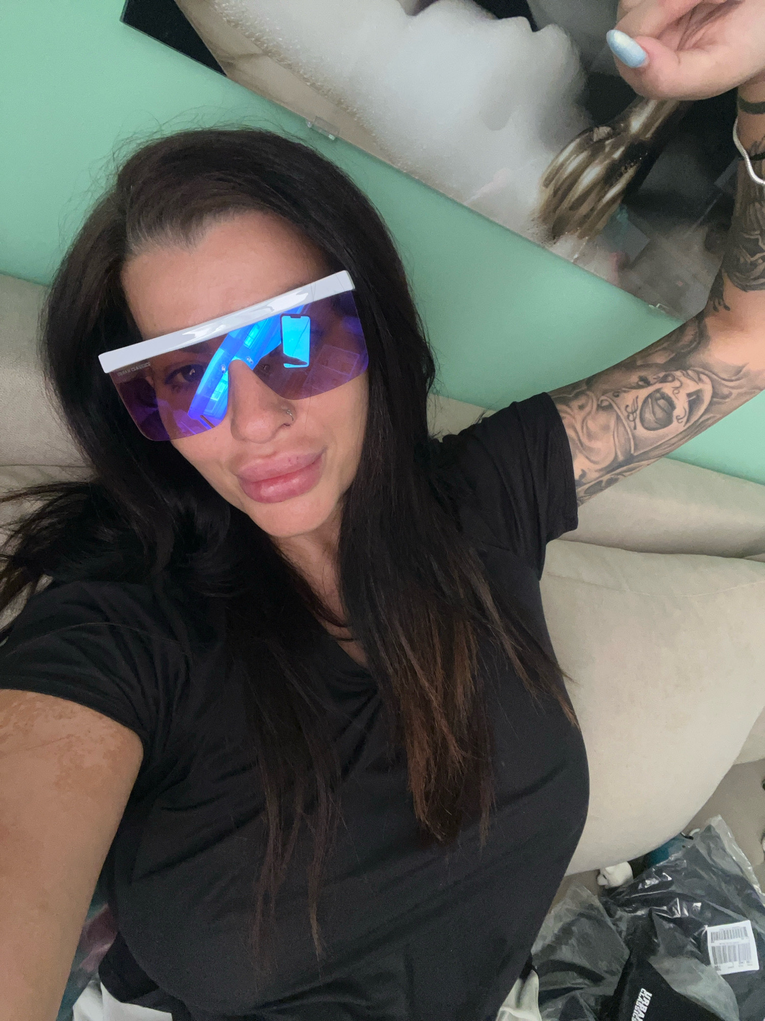 A woman with long, dark hair and large, reflective sunglasses is lying down, looking at the camera. She is wearing a black t-shirt. Her left arm is visible, showing a tattoo. Her right arm is raised, and her hand is near her face. The background appears to be a couch or bed with a mint green wall behind it. There are some folded items of clothing in plastic packaging on the right side of the image. No specific location in Plovdiv, Bulgaria, can be identified from the image.