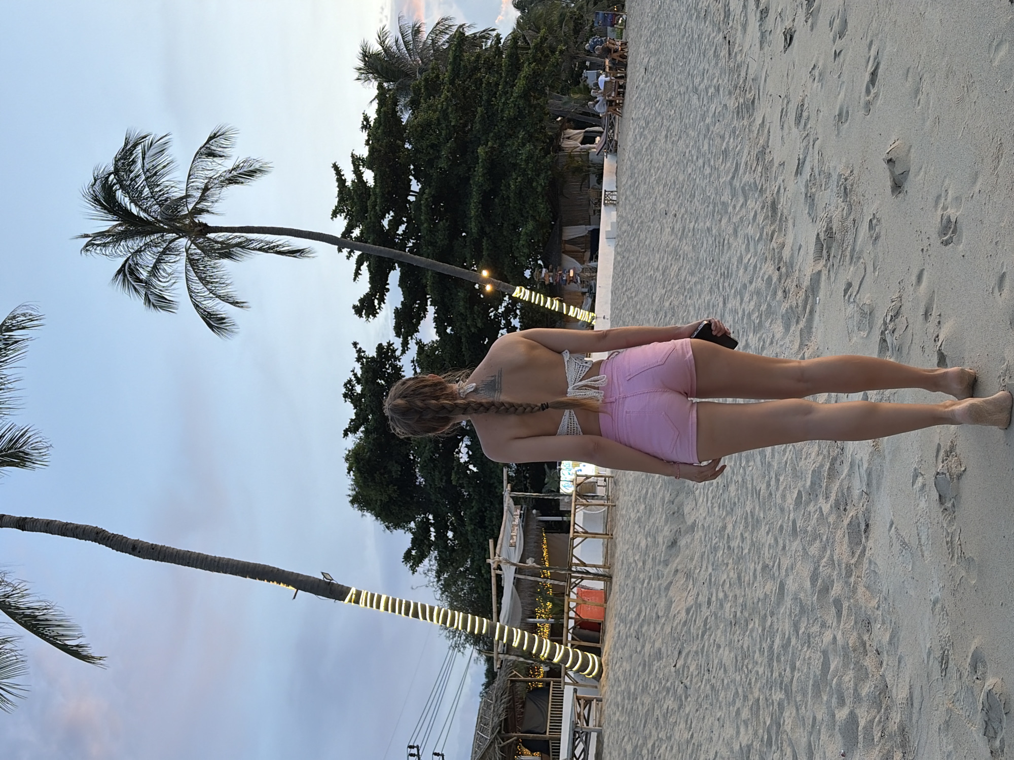 A woman with long blonde hair in a braid, wearing a white crocheted top and pink denim shorts, stands on a sandy beach facing away from the camera. She is holding a dark-colored smartphone in her right hand. The scene is set on a beach with palm trees, some decorated with strings of yellow lights. In the background, there are several open-air structures, possibly restaurants or bars, with thatched roofs and bamboo elements. People are seated at tables in the background. The sky is a dusky blue with hints of pink, suggesting it is either sunrise or sunset. The sand is light-colored and shows footprints. The overall atmosphere appears relaxed and tropical.