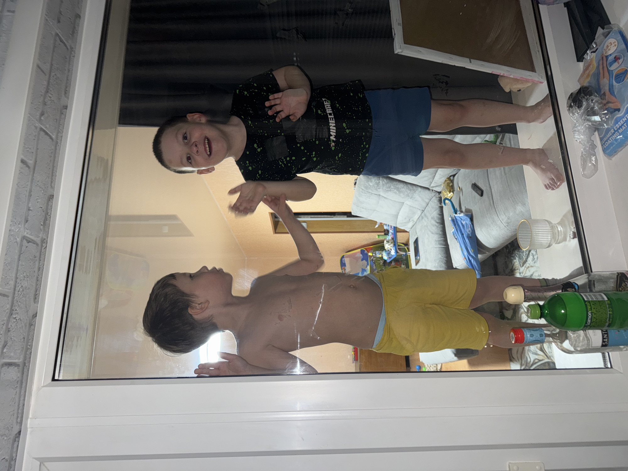 A photograph shows two children reflected in a window. One child is wearing a dark t-shirt with "Minecraft" written on it and blue shorts. The other child is wearing yellow shorts and is shirtless. The reflection also shows the interior of a room with various objects, including bottles, glassware, and a floating mat. The photo was taken in Chaikovsky, Russia.