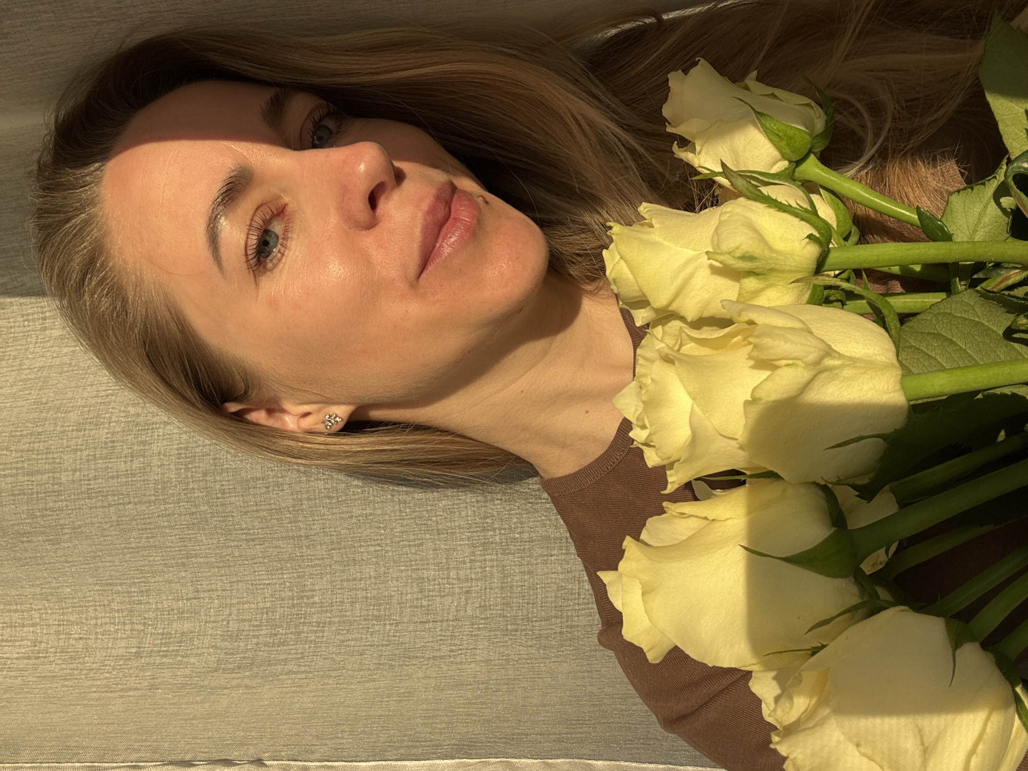 A light-skinned woman with blonde hair lies on a textured surface, likely fabric, in a supine position. She is wearing a brown top. Her head is turned towards the camera, and her eyes are open. Next to her, on her right, are a bouquet of yellow roses with visible green stems and leaves. The lighting suggests an indoor setting. The location is presumably Chaikovsky, Russia, but there are no elements in the image to confirm this.