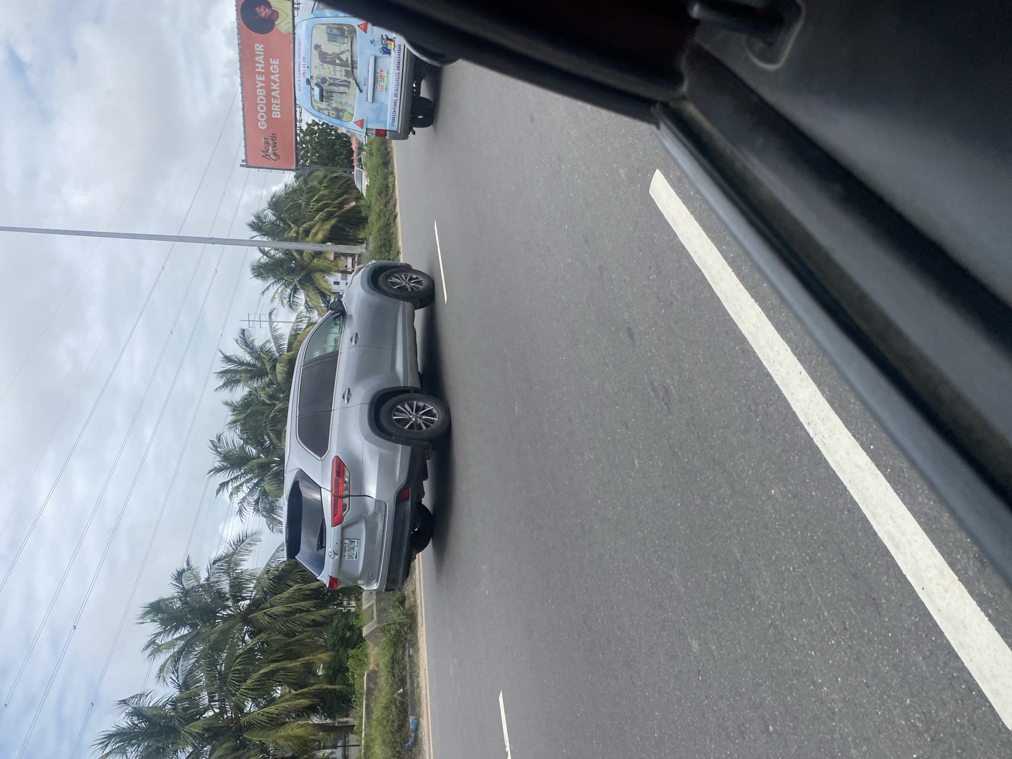A silver Toyota Highlander SUV is captured from a low-angle, slightly elevated perspective, driving on a paved road with a white line marking the lane. The vehicle's rear is facing the camera. The SUV has a license plate that reads "AKD 942 HW". In the background, palm trees line the side of the road, with power lines stretching across the cloudy sky. To the right of the SUV, a light blue van is visible, also in motion. The van has graphics on its rear depicting people cleaning and a phone number. A billboard with the text "Mega Growth GOODBYE HAIR BREAKAGE" is visible behind the van. The scene appears to be in Ilado, Nigeria, given the vegetation and the style of vehicles and billboards. The perspective suggests the photo was taken from another vehicle.