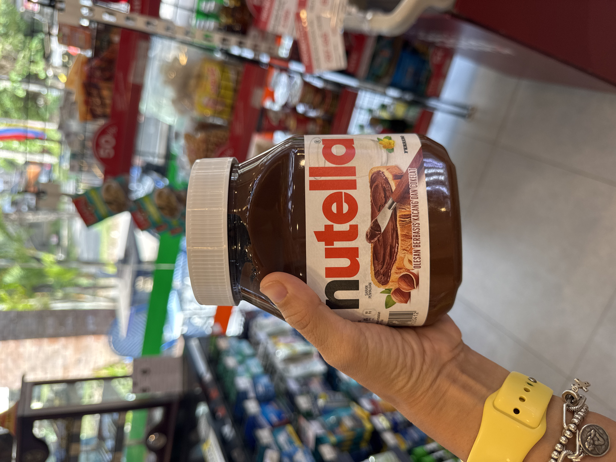A hand wearing a yellow wristwatch and silver charm bracelet is holding a jar of Nutella in a store, presumably in Kuta Selatan, Indonesia. The label on the Nutella jar is in Indonesian. Rows of packaged products are visible on the shelves behind the hand. Reflections from the store's exterior are visible on the glass shelving.