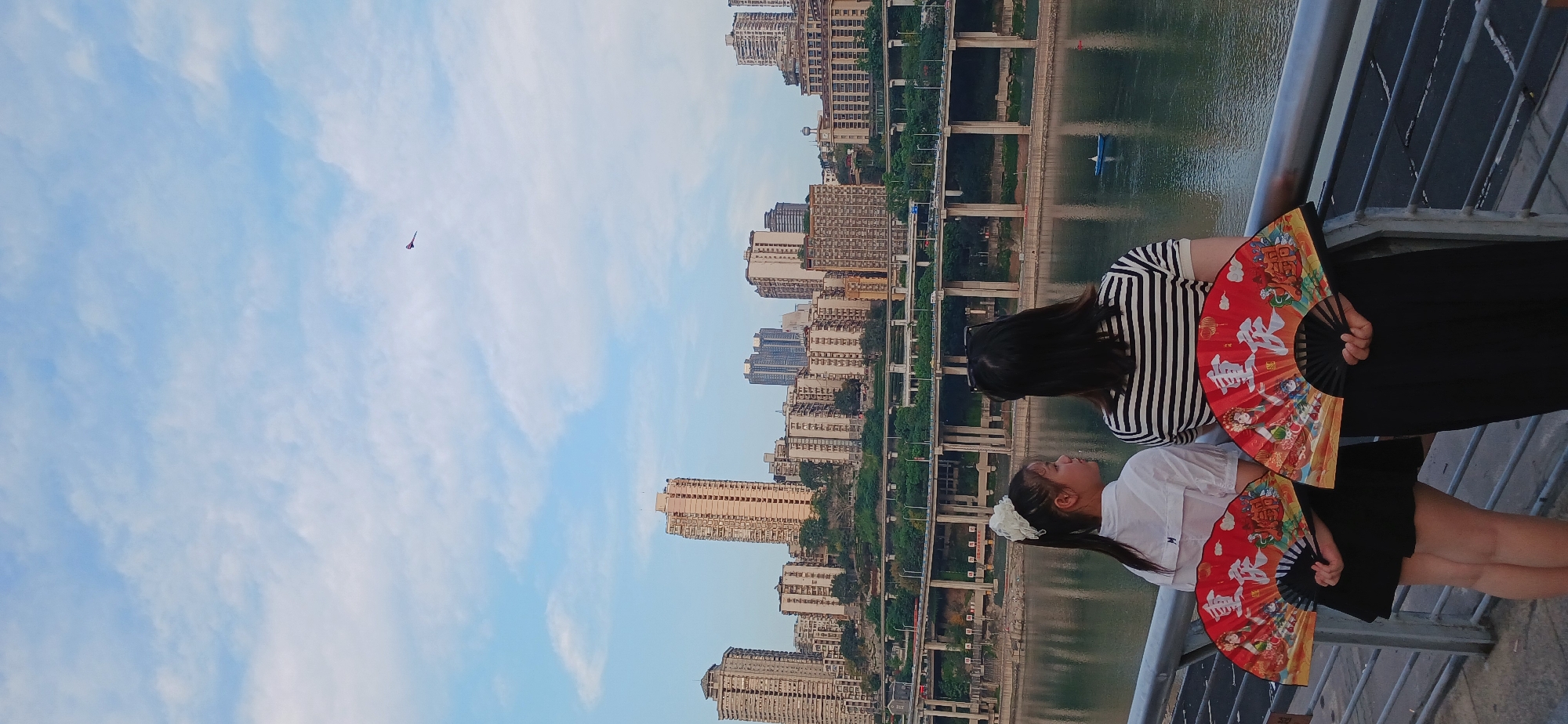 Two females stand on a bridge in Yuzhong District, China, holding red hand fans. The background shows urban buildings, bridges, and water. The sky is partly cloudy.