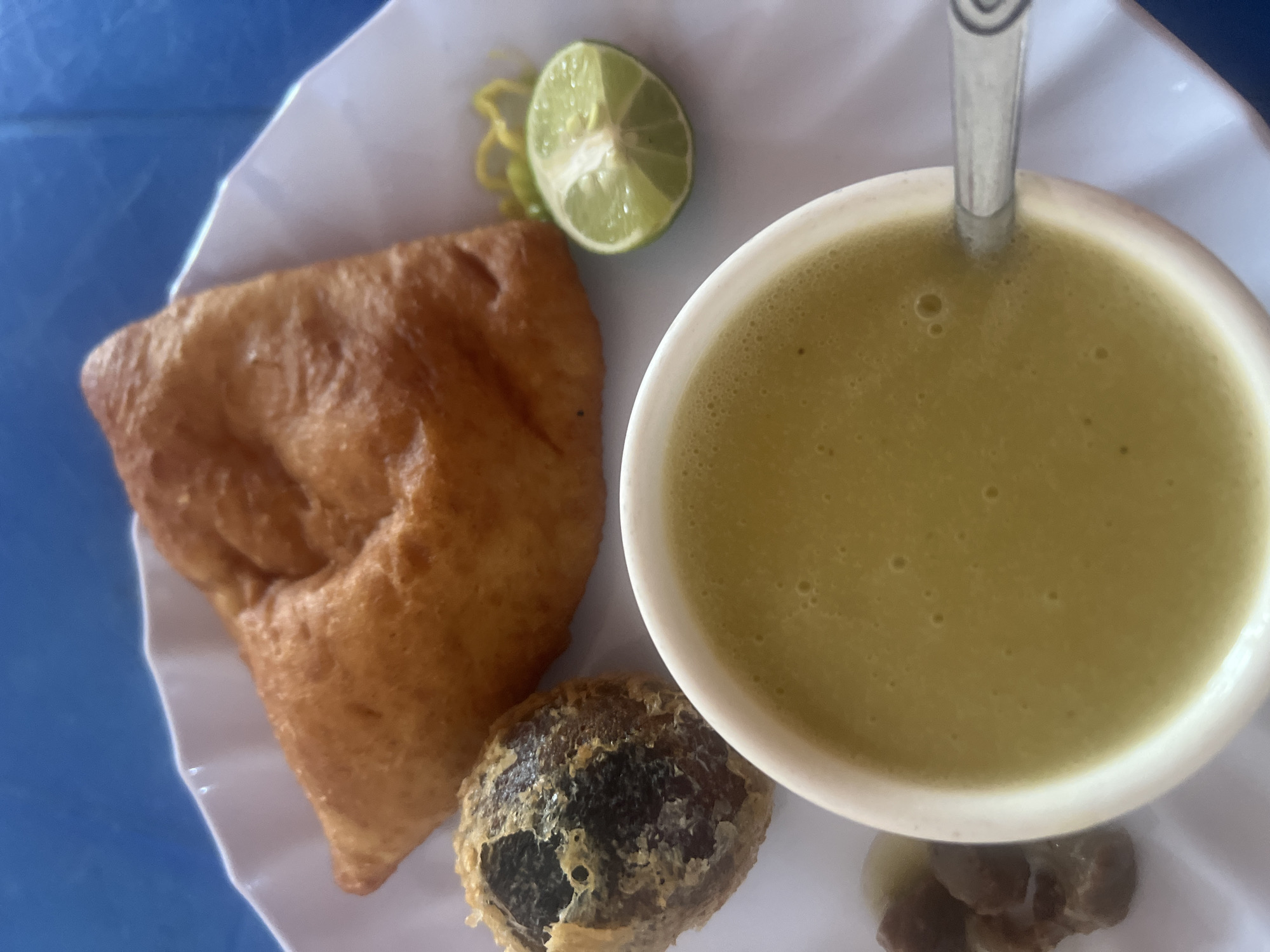 The image shows a meal on a white plate. The meal consists of a large, golden-brown fried dough item, a fried ball with a dark interior, a slice of lime, and a white bowl filled with a green liquid, possibly soup. A spoon is in the bowl. There are also some small pieces of meat or vegetables on the plate. The plate is placed on a blue surface. This meal is likely being served in Dar es-Salaam, Tanzania.