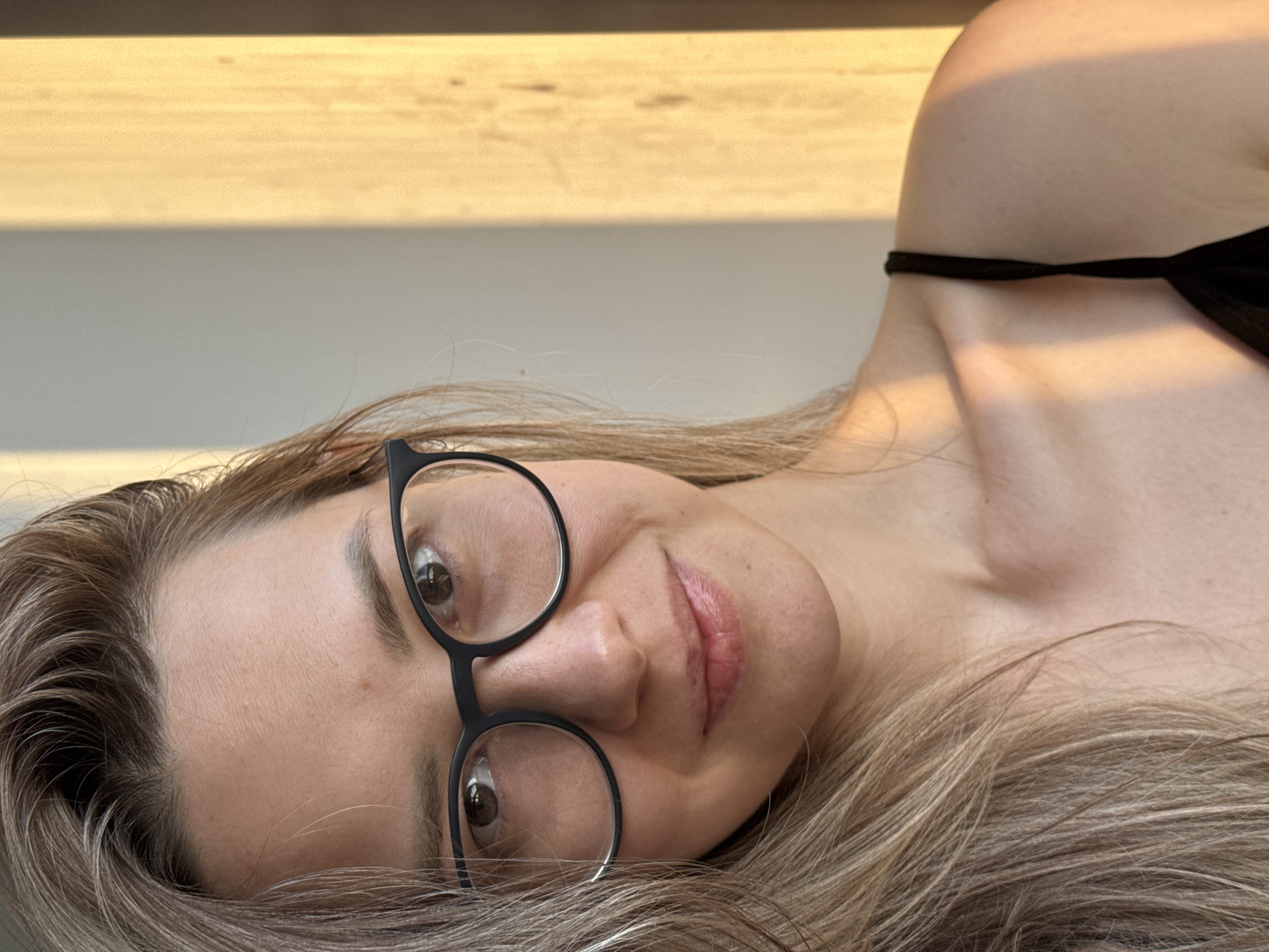 A woman lies on her side. She is wearing glasses and a black strap is visible. The background includes a light brown, horizontal surface. There is no indication this photo was taken in Chaikovsky, Russia.