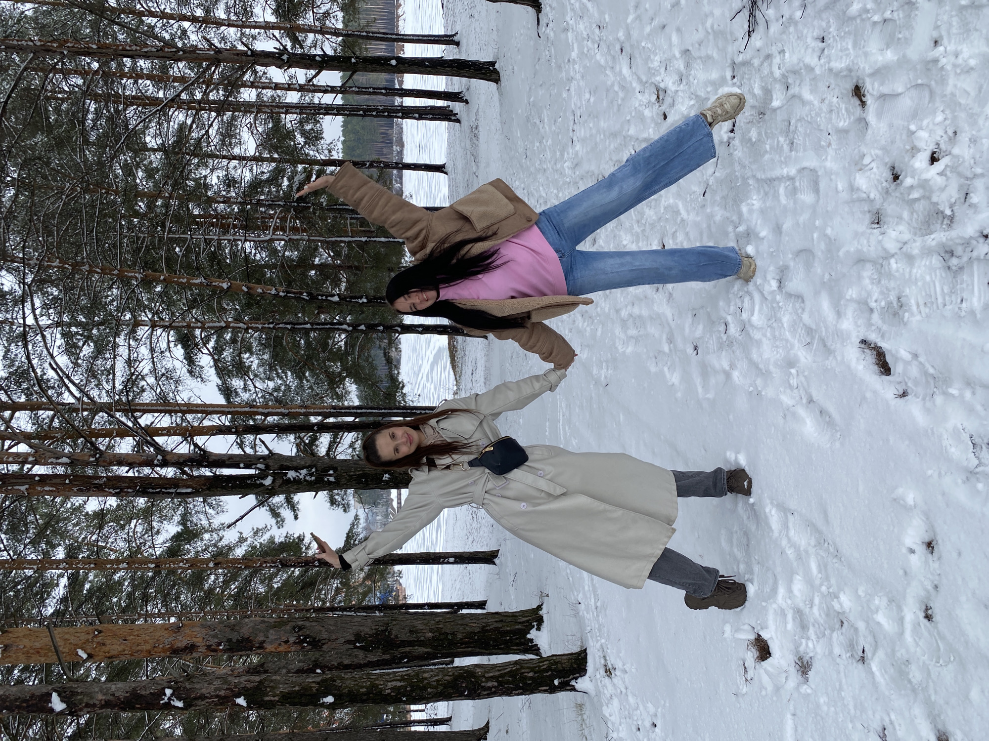 Two women are lying head-to-head in the snow near a forest in Chaikovsky, Russia. The woman on the left wears a long, light-colored trench coat with a black waist bag, dark gray pants, and dark boots. She extends both arms upwards. The woman on the right wears a brown jacket over a pink sweater, blue jeans, and light-colored sneakers. She also has both arms extended upwards. There are tall pine trees on either side of the women, and a body of water is visible behind the trees in the background. The ground is covered in snow.