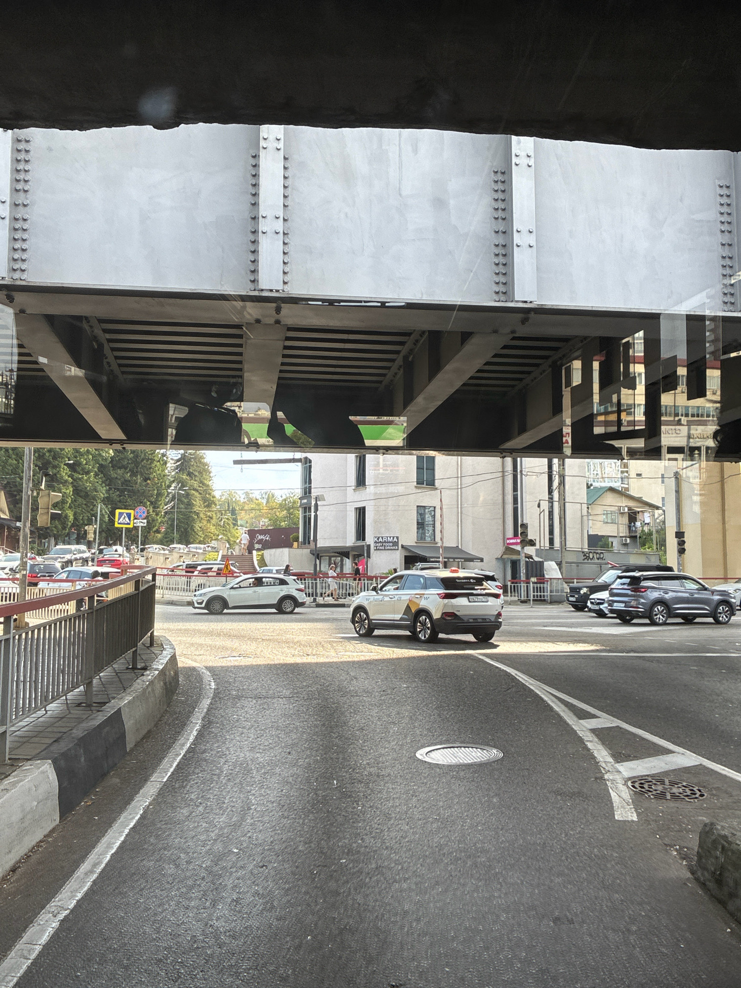 The image captures a view of a city street in Sochi, Russia, from beneath a large metal bridge structure. The perspective is from a vehicle, looking forward and slightly downwards. The foreground shows a section of asphalt road with white lane markings and two manhole covers. To the left, there is a concrete barrier with a metal fence on top, bordered by a curb with black and grey paint.
In the mid-ground, several cars are visible on the road, including a white SUV and a darker SUV. The white SUV has a taxi sign on its roof. A pedestrian crossing sign with a yellow background and a blue pedestrian symbol is visible further down the road. Buildings with white facades and windows are seen on the right side of the street. Trees line the left side of the road.
The background shows more buildings and more trees. The sky is clear and bright, suggesting daytime. Reflections of buildings and the sky are visible on the underside of the bridge structure, which is made of large metal beams and plates connected with rivets. The overall scene depicts an urban environment with traffic and infrastructure.