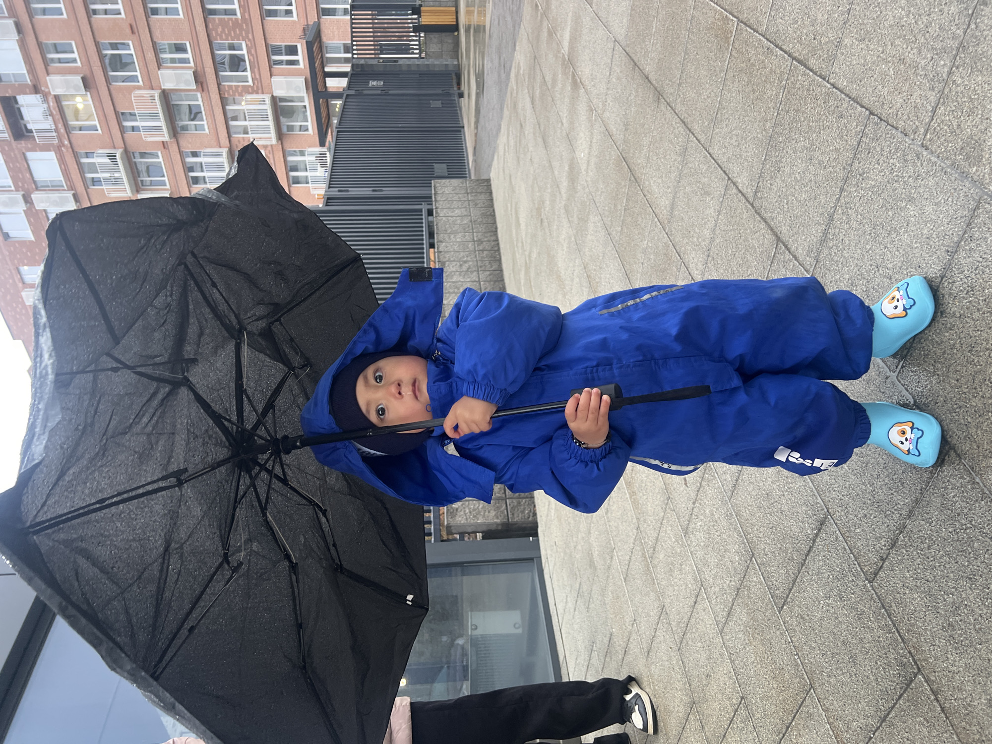 An image, captured in Tyumen, Russia, depicts a young child dressed in a bright blue waterproof snowsuit or rainsuit, standing outdoors on what appears to be a wet, overcast day. The child, likely a toddler, is positioned centrally, holding the handle of a large black umbrella, which is open and visibly covered in water droplets. The umbrella is held partly by the child and partly by an adult whose legs and black sneakers are partially visible in the bottom left of the frame.
The child is wearing a dark blue beanie under the hood of their suit and light blue rain boots adorned with cartoon dog faces on the toes – one boot features a dog with a blue hat/scarf, the other shows two smaller dogs. The child's face is turned towards the camera, with wide, curious eyes.
The setting is an urban outdoor space with grey, rectangular paved ground that appears damp, suggesting recent rain. In the background, tall, multi-story brick buildings with numerous windows and external air conditioning units are visible, characteristic of city architecture. Dark metal fences or railings run along the base of some of these structures. The light suggests it is daytime, but the overcast sky indicates a cloudy or rainy condition. There is some indistinct white text or a logo on the lower leg of the child's blue suit, but it is not clearly legible. The overall mood is one of a child exploring the elements on a rainy day, perhaps under the watchful eye of an adult.