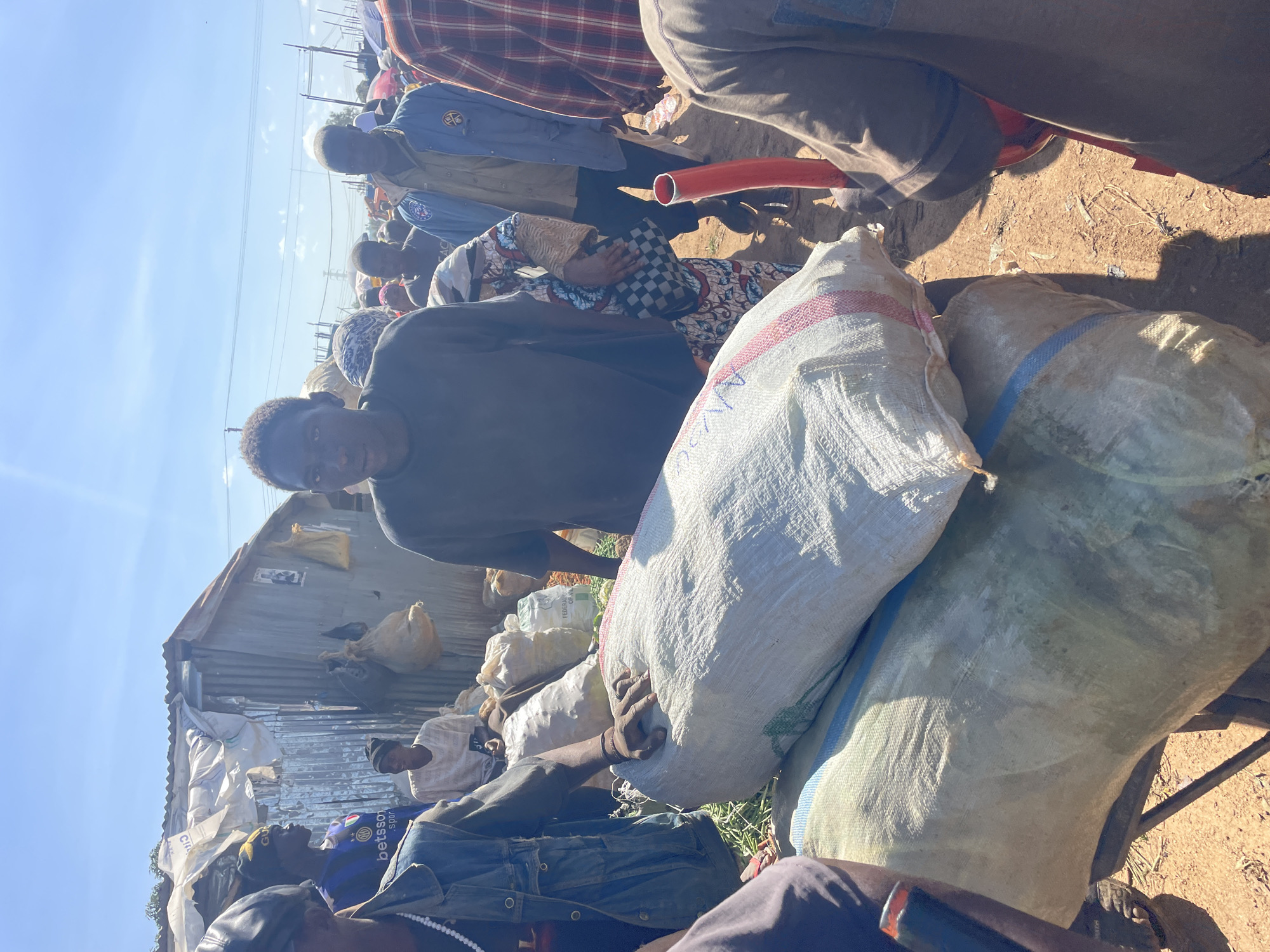 The image captures a bustling outdoor market scene in Gura Top, Nigeria, likely in the afternoon given the bright sunlight and clear sky. A group of people are gathered around large sacks, possibly agricultural produce or goods for sale. In the foreground, two large, light-colored sacks are prominently displayed, one stacked on top of the other. The upper sack has red and white stripes running horizontally.
Several individuals are visible. A young man in a dark t-shirt is looking directly at the camera, positioned in the center of the frame. To his left, a person wearing a blue denim jacket and a black cap stands, with a white beaded necklace visible. Other individuals are scattered throughout the scene, some in the background appearing to interact with vendors or browse goods. One person is wearing a blue jacket with a logo on the left breast. A woman in the background is wearing a patterned dress and holding a checkered bag.
The environment appears to be a somewhat informal market setting. In the background, a simple structure with a corrugated metal roof and siding, possibly a makeshift stall or dwelling, is visible. Other market stalls with umbrellas can be seen further in the distance, indicating a lively commercial area. The ground is dusty, typical of an outdoor market. A red pipe-like object is held by someone in the lower right portion of the image. The overall impression is one of everyday commerce and community activity in an urban Nigerian setting.