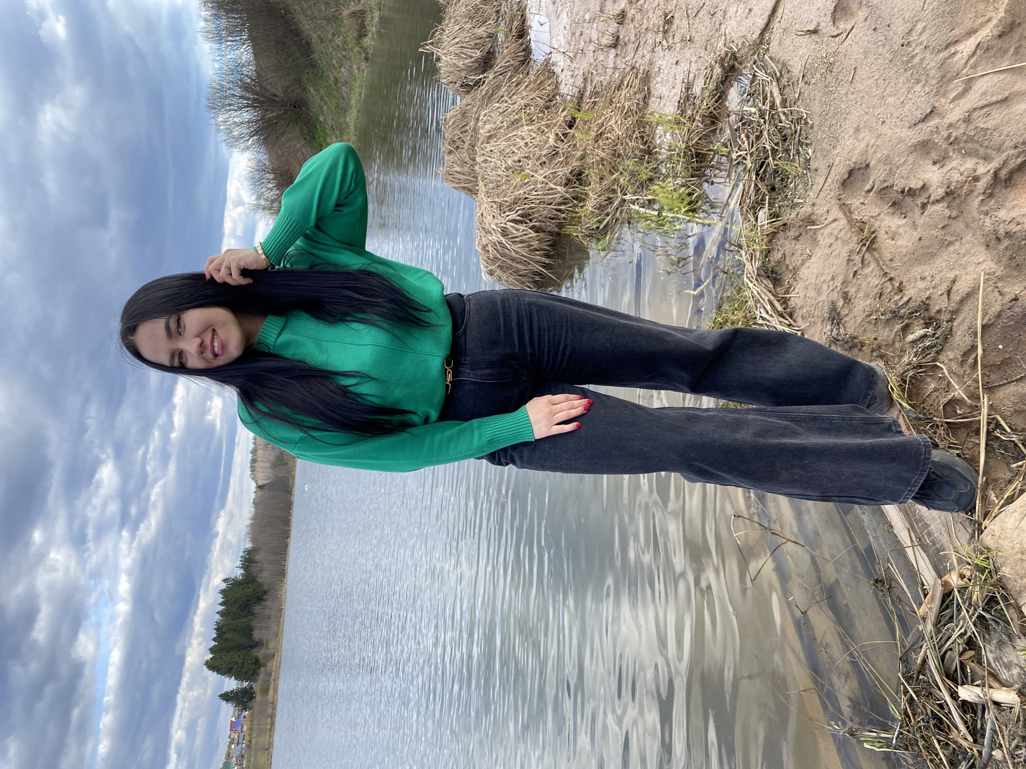 A young woman is captured in a full-length portrait, standing alongside a body of water. She has long, dark hair and is wearing a bright green long-sleeved sweater, dark grey or black wide-leg pants, and dark shoes. She is smiling genuinely at the camera, with her right hand casually resting near her head, showcasing a gold-colored bracelet on her wrist. Her left hand, with red nail polish, is lightly touching her hip.
She stands on a muddy bank adorned with dry grasses and reeds. The water surface is calm, reflecting the cloudy sky above. In the background, across the water, a horizon line features a dense treeline composed of both bare deciduous trees and green pine trees, hinting at the early spring season. Further in the distance, a few small houses or buildings are discernible. The sky is predominantly overcast with varied shades of grey clouds. The overall scene suggests a rural or semi-rural setting in Foki, Russia. There is no visible text in the image.