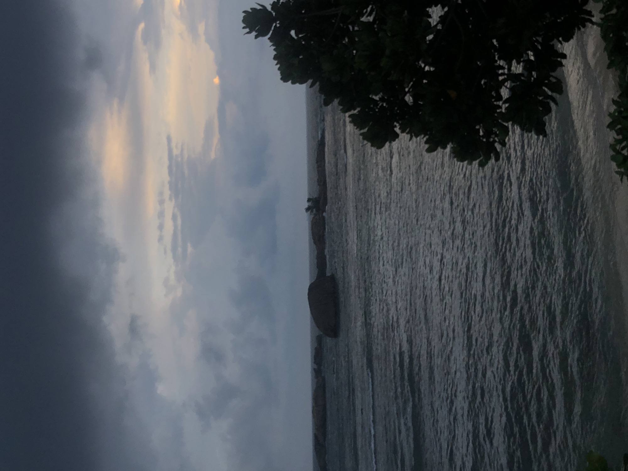 The image captures a view of the ocean from a coastal location in Galle, Sri Lanka. The sky is overcast with dark storm clouds on the left, contrasting with lighter, sunlit patches on the right. The water is dark and appears slightly turbulent. The shoreline is rocky and lined with some vegetation including trees in the foreground that are partially obscuring the view.