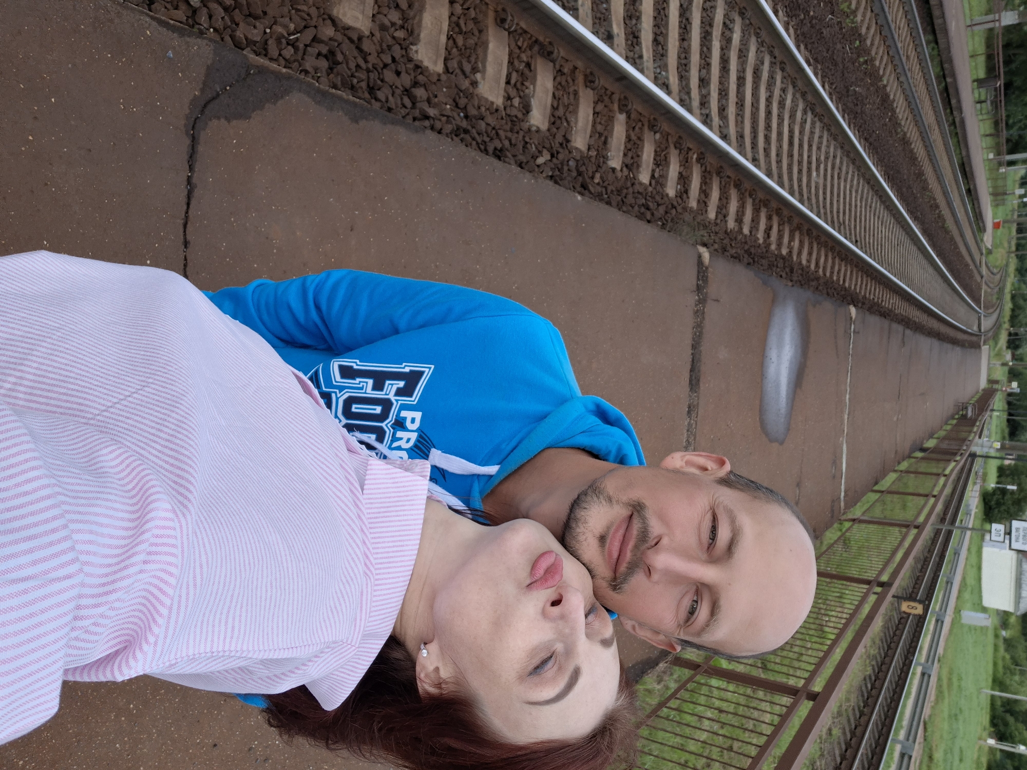 An outdoor, waist-up, selfie-style, high-angle shot shows a bald, middle-aged man with a goatee and a woman with auburn hair, both posing on a train platform. The man, on the left and slightly behind the woman, wears a blue hoodie with white text. The woman, on the right and in front, has her eyes closed and her lips pursed in a kiss, and wears a pink and white striped shirt. They are positioned in the foreground, with train tracks and a platform extending into the background. The environment appears to be a rural train station, possibly in Забалоцкі сельскі Савет, Belarus, given the presence of what appears to be Cyrillic signage in the distance. The platform is made of concrete, and there are metal railings and grass visible. The sky is overcast.