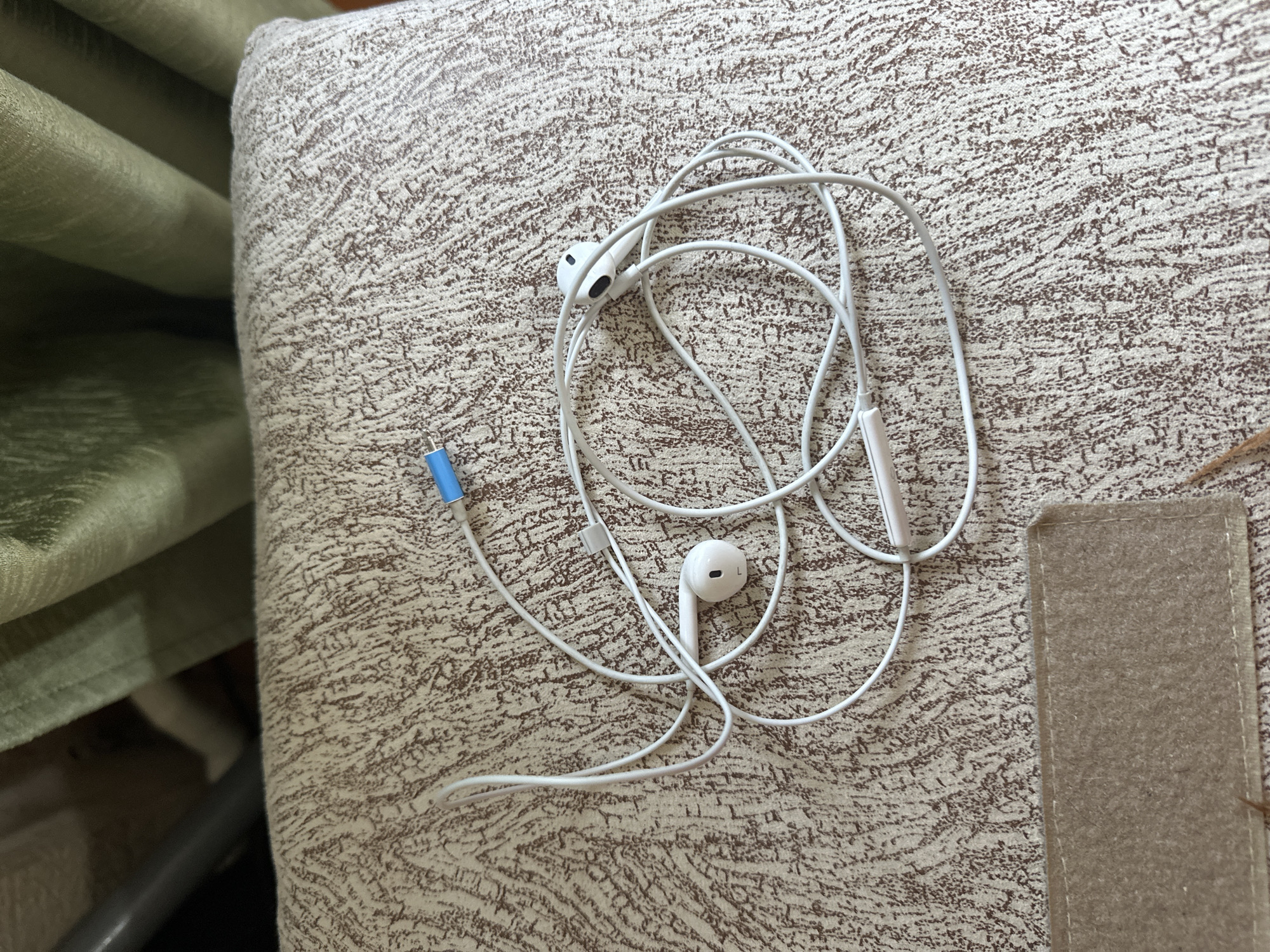 A tangled pair of white wired earbuds with a blue connector lies on a textured, beige and brown patterned fabric surface, likely a sofa or cushion. The earbuds are coiled and spread out, with one earbud clearly visible on the left and another on the right. A grey tube and green draped fabric are visible in the background. The image was taken indoors, likely in a residential setting in Rostov-on-Don, Russia, judging by the provided metadata. There are no people or animals present in the image.