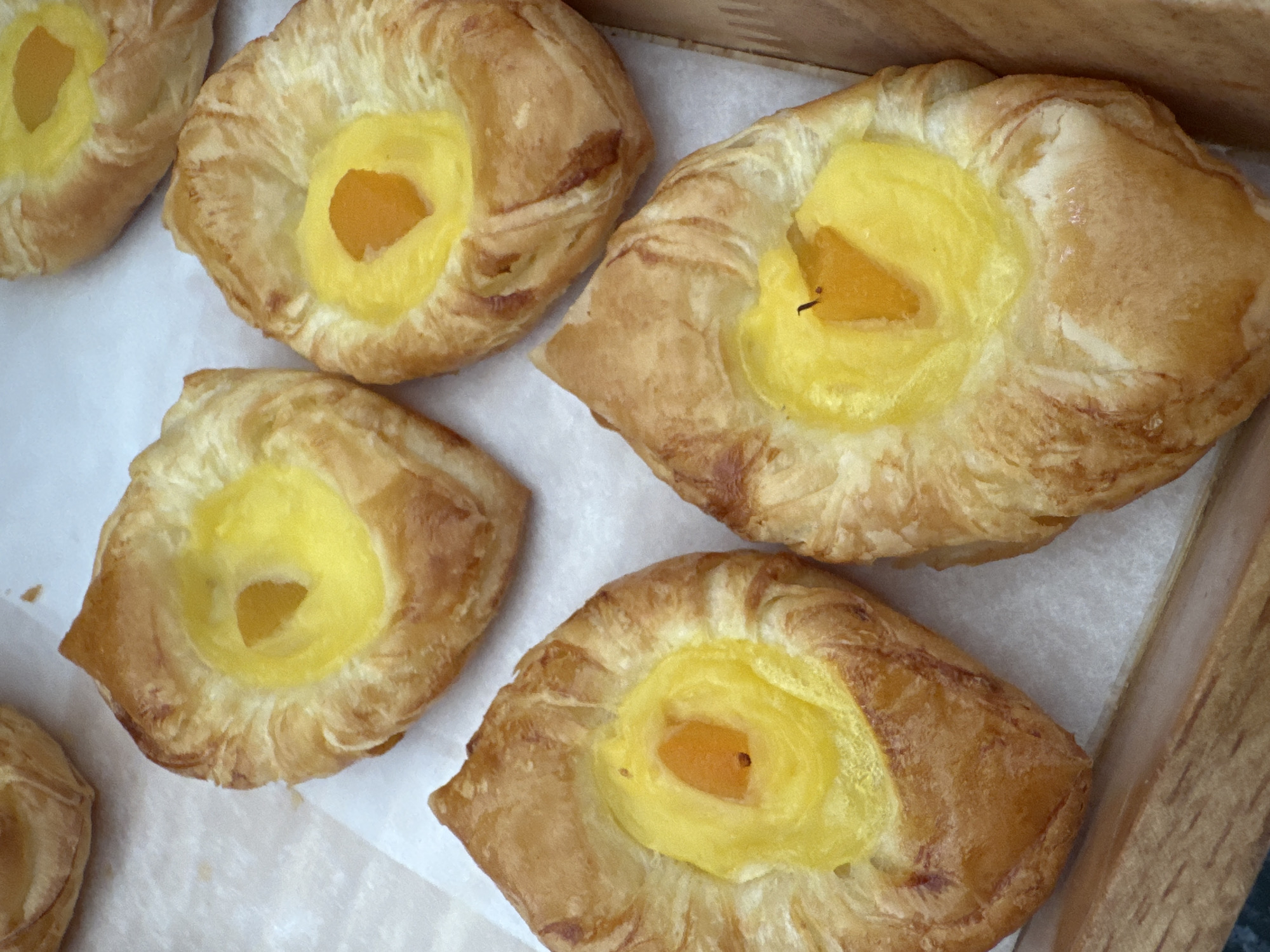 The image displays a close-up, overhead view of six golden-brown pastries, likely Danish or puff pastry, arranged on a sheet of white parchment paper inside a light brown, possibly wooden or cardboard, tray. The scene is set in a commercial or domestic kitchen environment within Nha Trang Ward, Vietnam. Each pastry features a central depression filled with a bright yellow custard or pastry cream. Embedded within the yellow filling of each pastry is a single, irregularly shaped piece of orange-colored fruit, consistent with an apricot or peach segment. Small, dark, linear elements, possibly remnants of stems or baking residue, are visible on the fruit pieces in some pastries. The pastries exhibit a flaky texture on their crusts, indicating lamination. The lighting is bright and even, illuminating the details and colors of the baked goods. No human subjects or additional environmental details are present.