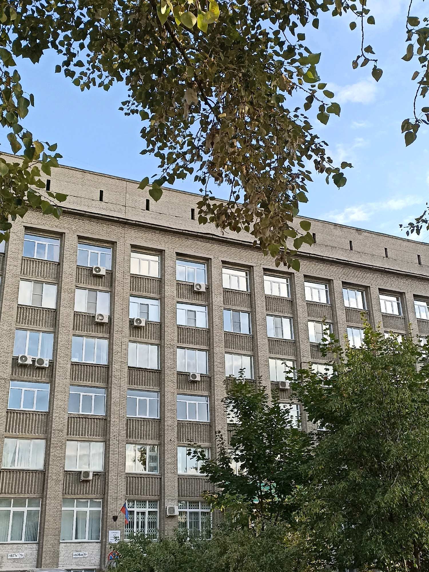 The image shows the facade of a large, light-brown brick building with multiple rows of windows. Trees with green leaves frame the upper and lower portions of the building. The sky above is blue with scattered white clouds. The building features numerous air conditioning units mounted on the exterior walls. Some windows are partially obscured by decorative grates or blinds. A small flag and some signage are visible near the ground level entrance. The architecture suggests it is likely an institutional or administrative building.