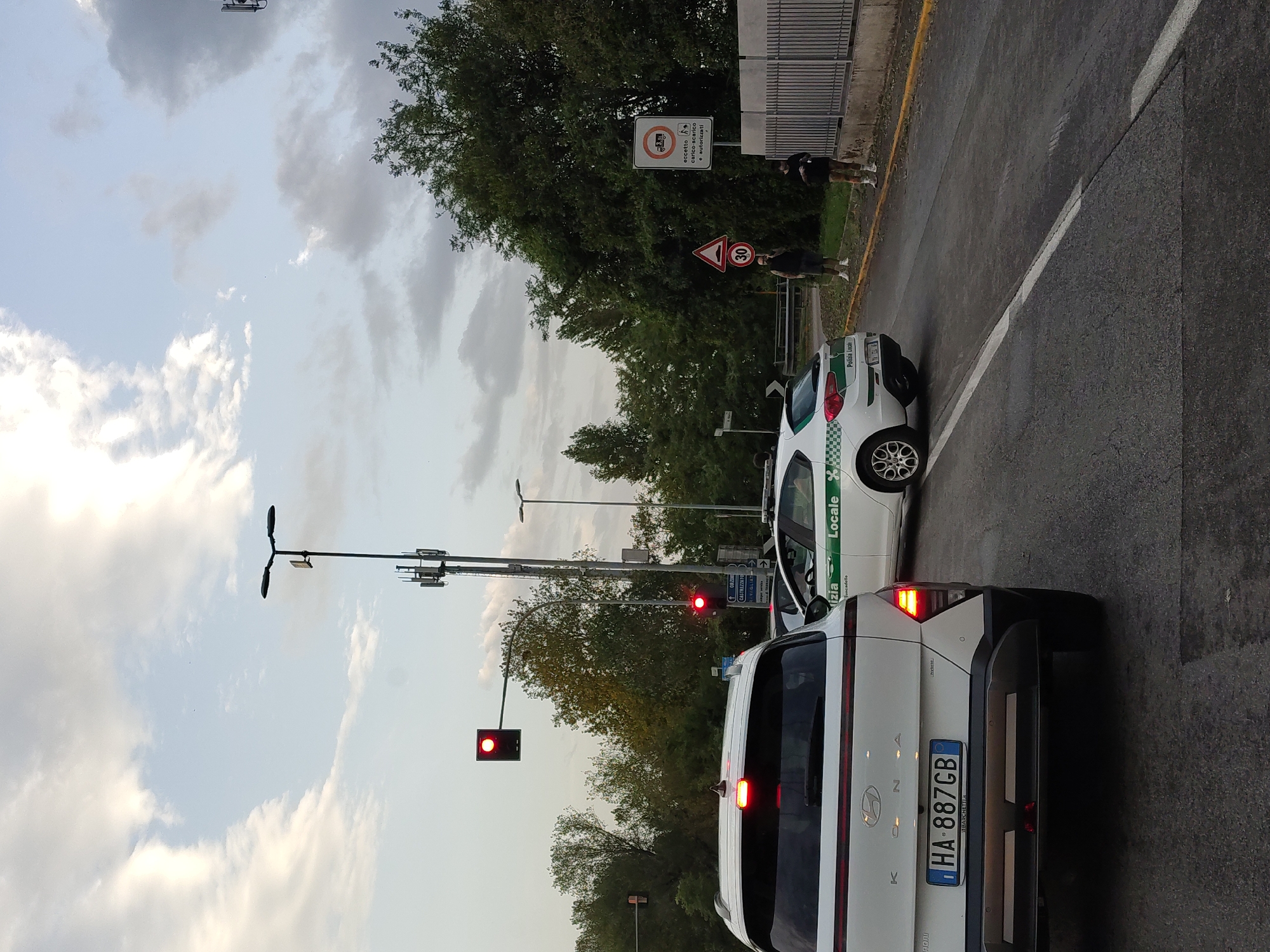 The image depicts a street scene in a city with two police cars and two civilians. The sky is overcast, with clouds suggesting it might be late afternoon or evening.
In the foreground, the rear of a white Hyundai Kona is visible, displaying an Italian license plate reading "HA 887GB". To its right, a white police car with the text "Polizia Locale" is parked, partially obscuring the view of a second white police car further down the road.
Two civilians are standing on the sidewalk to the right of the road. One is a male wearing a dark t-shirt and shorts, holding a mask. The other is a female wearing a black t-shirt and shorts, with her hair in a bun. Both are facing away from the camera and towards the street.
Traffic lights are visible, with at least one showing red. Road signs include a speed limit sign indicating 30 and a sign with a symbol of a truck and a line through it, suggesting a restriction for trucks. Trees line the right side of the road, and a metal fence runs along the sidewalk. The road surface is asphalt, with visible lane markings.
