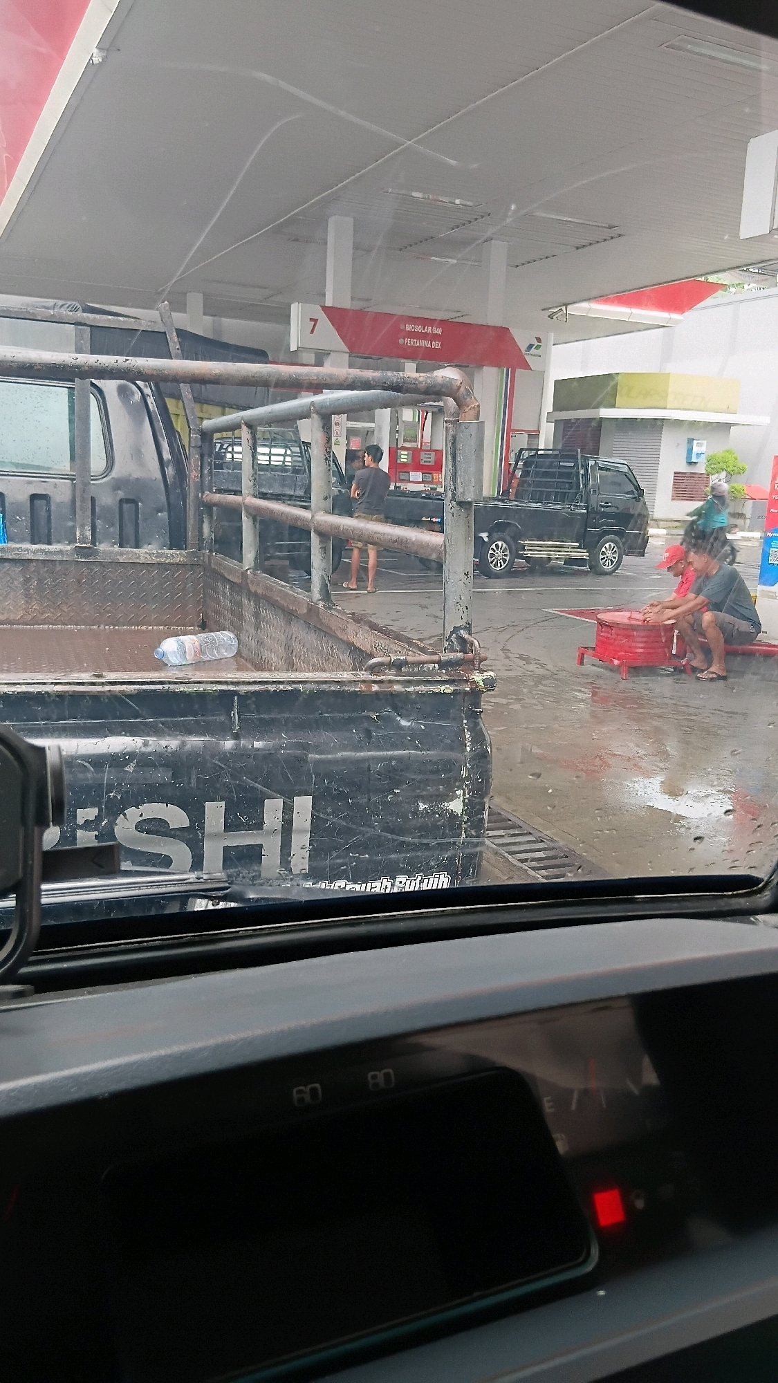 The image captures a scene at a Pertamina gas station in Batusangkar, Indonesia, viewed from inside a vehicle. In the foreground, the back of a black pickup truck is visible, with its bed containing a water bottle and some debris. The truck has "MITS" partially visible on its tailgate. Beyond the truck, at gas pump number 7, two individuals are seated on a red circular object. One is wearing a red cap and a dark shirt, while the other wears a dark t-shirt and shorts. They appear to be interacting or resting. Another black pickup truck is parked at a gas pump in the middle ground. Several other people are present, including two individuals standing near a gas pump and another person walking in the background. The ground is wet, indicating recent rain or cleaning. The gas station canopy is visible overhead, with red and white signage. The dashboard of the vehicle from which the photo is taken is partially visible in the bottom of the frame, showing the speedometer with markings at 60 and 80, and a fuel gauge with the needle pointing towards empty, a red warning light illuminated, and the letter "E" for empty.