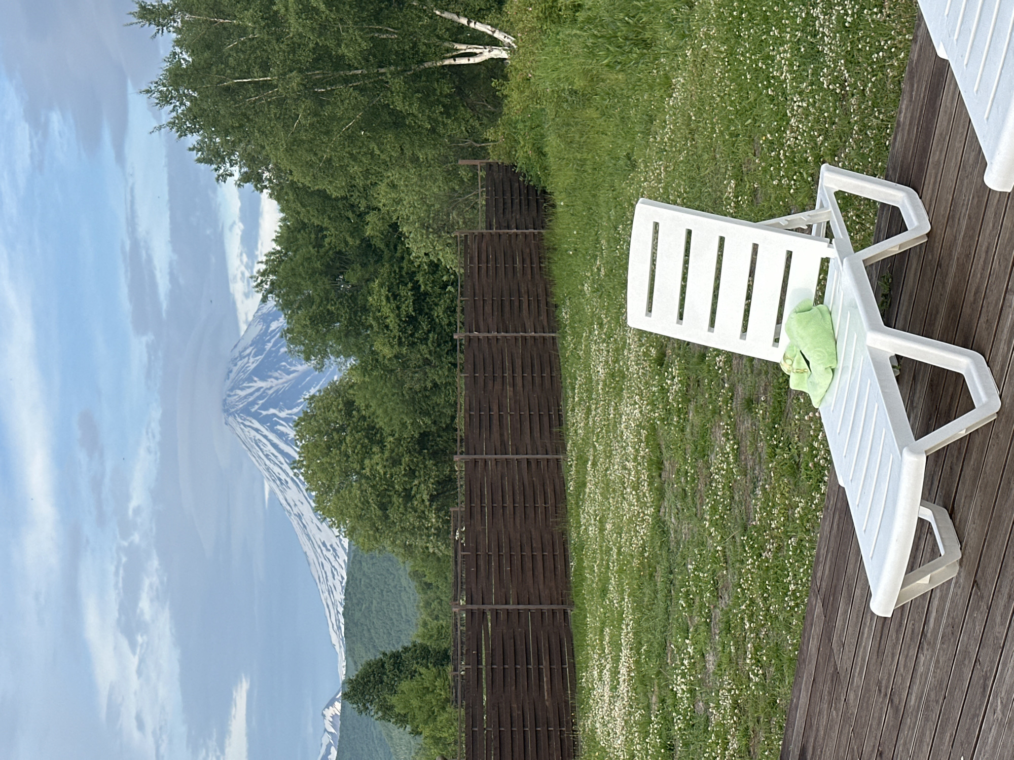 In Yuzhnyye Koryaki, Russia, a white plastic chaise lounge sits on a wooden deck. A green towel is draped over the lounge. Beyond the deck, a well-maintained lawn is dotted with small white flowers. A dark wooden fence runs horizontally across the middle ground, separating the lawn from a dense forest. In the background, a snow-capped volcano dominates the horizon under a cloudy sky. The environment appears serene and natural, with a hint of recreational setup.