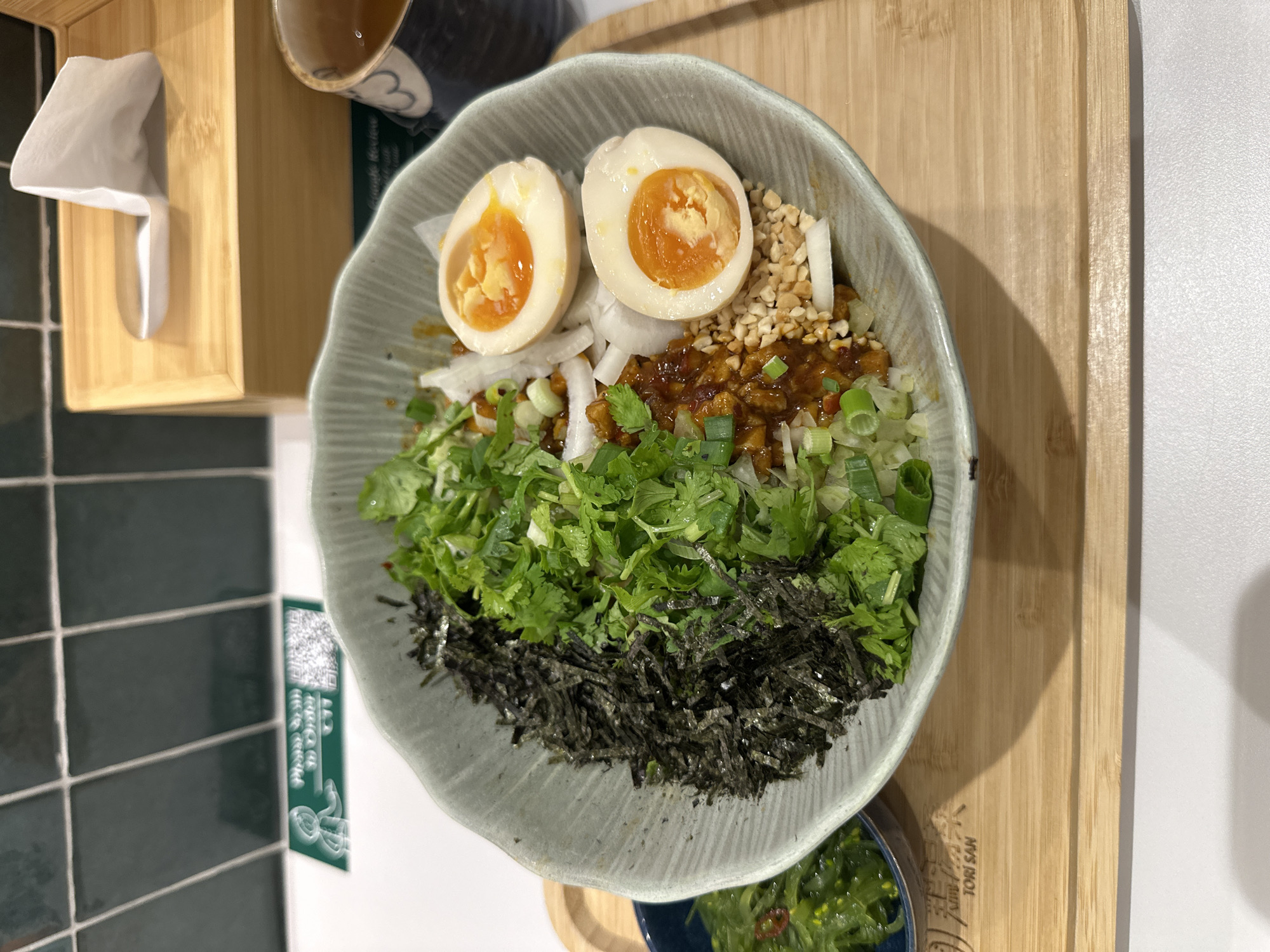 A close-up overhead shot shows a Japanese meal served in a light green, textured bowl with a wavy rim, set on a light wood tray. The bowl is filled with various ingredients, including sliced nori seaweed, chopped green onions, fresh cilantro, chopped peanuts, sliced white onions, a spicy brown sauce, and two halves of a soft-boiled egg with bright orange yolks. To the left of the main bowl, a small dark blue bowl contains a portion of green seaweed salad with small red chili pieces. In the background, a wooden tissue box dispenses a white tissue, and a dark blue and white patterned teacup with amber liquid sits to the right. The wall behind the food is tiled with dark green squares. The tray also has a logo with the text "TORI SAN" in English and Chinese characters, suggesting a location at a Japanese restaurant called Tori San in Singapore.