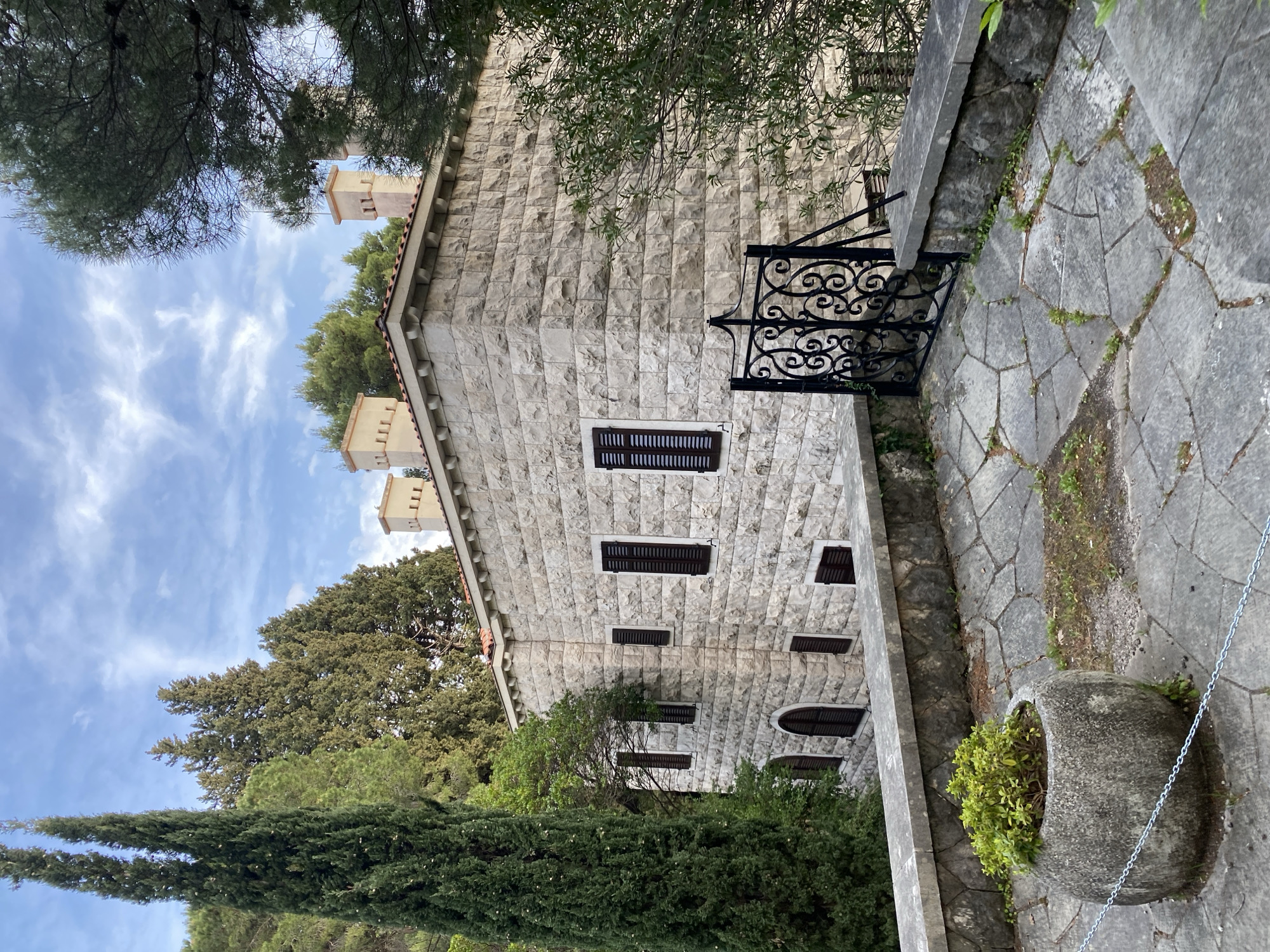 The image depicts a stone building in Sveti Stefan, Montenegro, surrounded by lush greenery. The building has a facade of light-colored stone blocks, with several windows adorned with dark shutters. A decorative black metal railing stands along the edge of a stone walkway, and a large stone planter with greenery sits nearby. The sky is blue with scattered clouds, and trees frame the building, suggesting a tranquil outdoor setting. The architecture and landscaping are characteristic of the Mediterranean region.