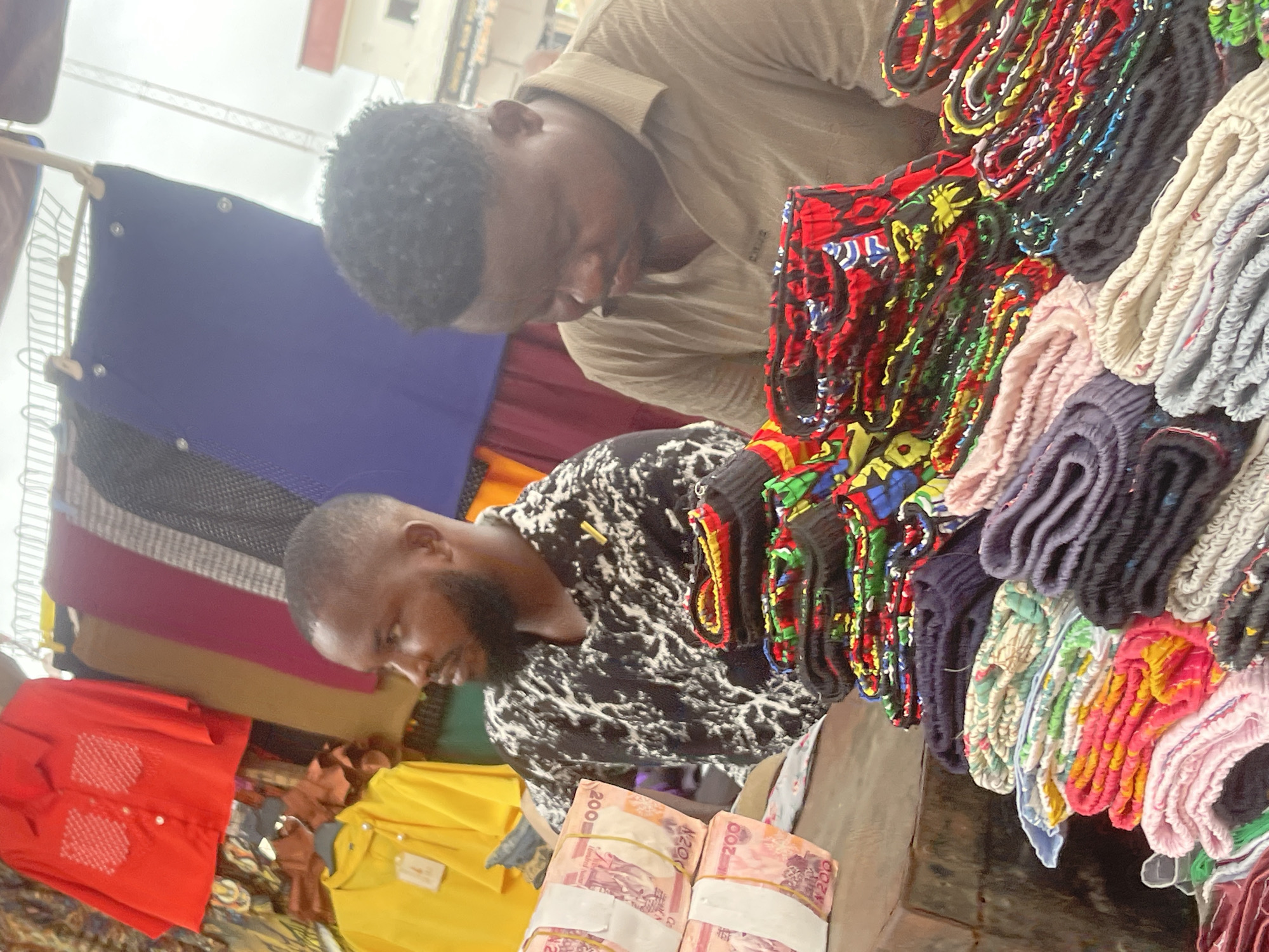 Two African men are engrossed in a transaction at a marketplace in Onitsha, Nigeria. One man wears a black and white patterned shirt, while the other sports a light brown shirt. They are positioned near stacks of brightly colored, patterned fabrics, possibly clothing items. Piles of banknotes, secured with rubber bands, sit on a surface near them, indicating payment. Assorted shirts in red and yellow are displayed nearby. The scene suggests a commercial exchange in a bustling market environment.