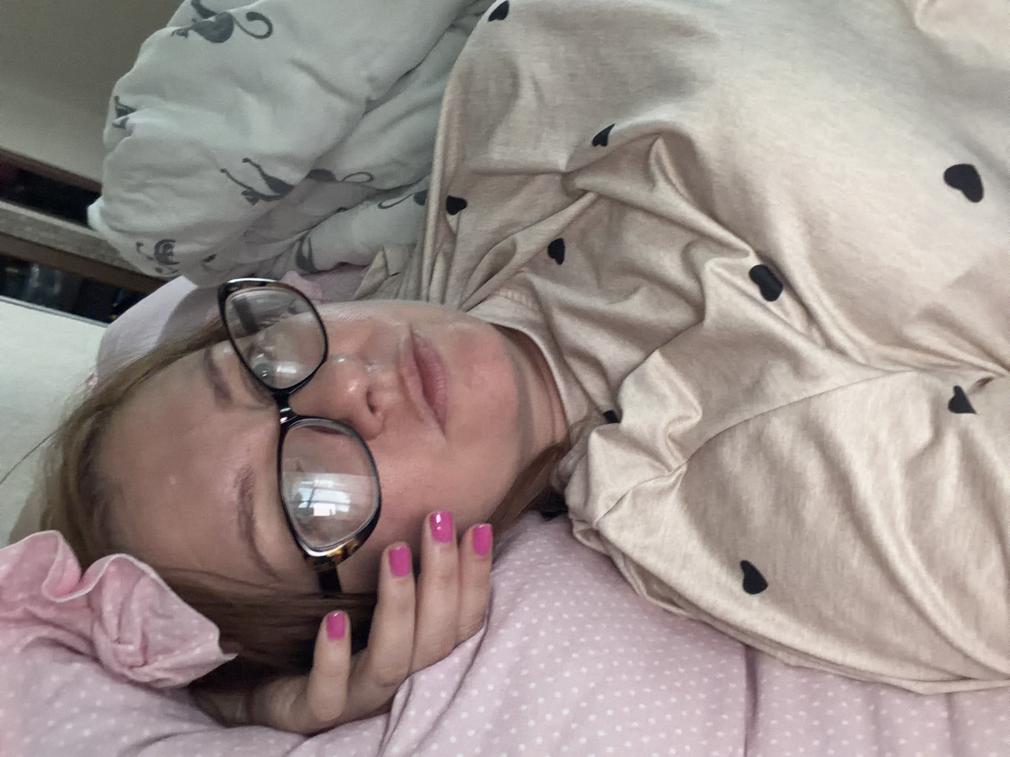 A woman with blonde hair and glasses lies in bed. She is wearing a light-colored shirt with black heart patterns. Her left hand, with bright pink fingernails, rests on a pink polka-dotted pillow. The background shows a white duvet with a gray cat pattern. The image is a selfie taken indoors. It is not possible to determine the specific location in Podolsk, Russia, from the image alone.