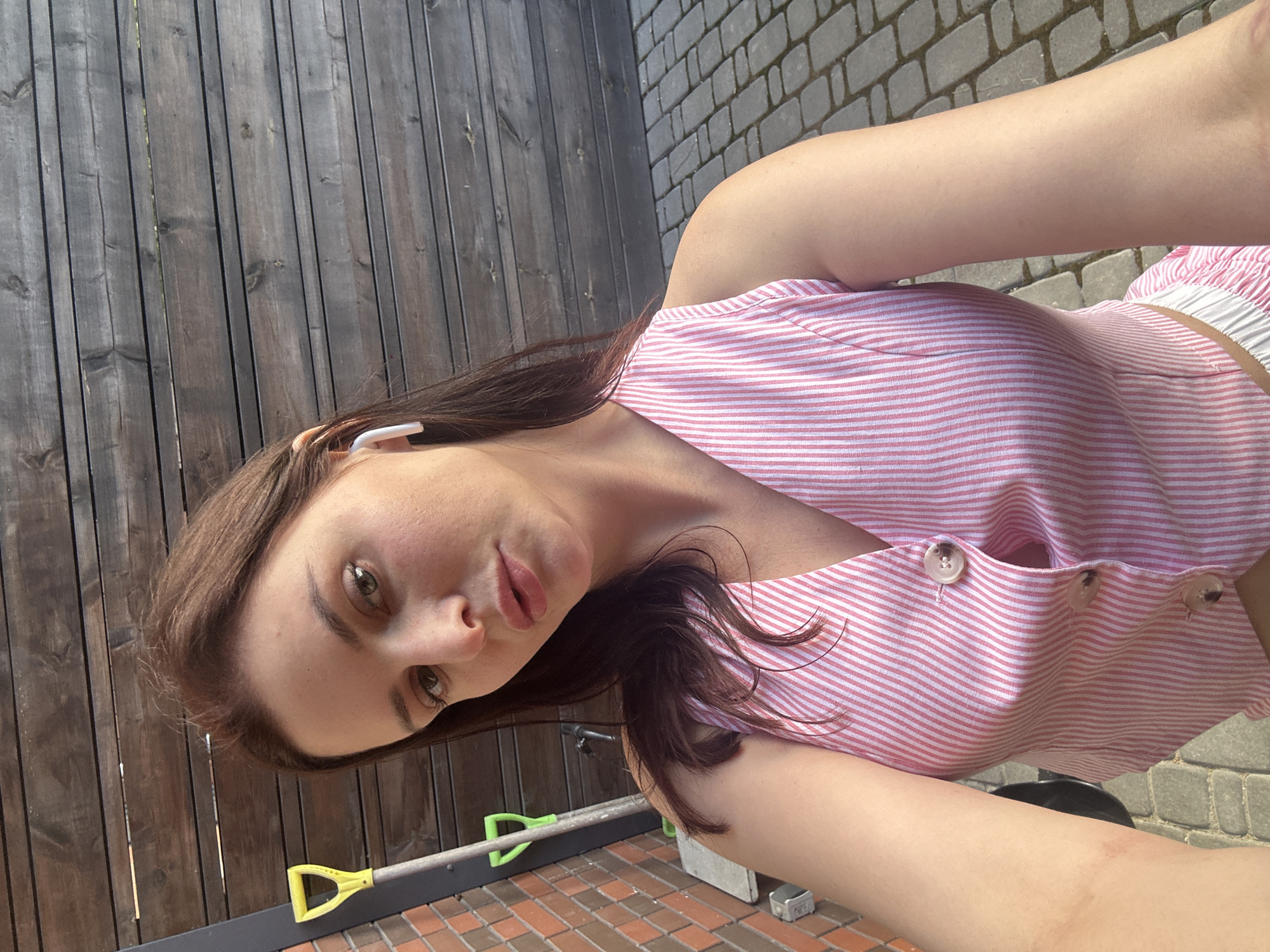 The image shows a young woman taking a selfie outdoors. She has long, dark brown hair and is wearing a pink and white striped crop top. She also appears to have a white earbud in her right ear. The background consists of a wooden fence and a paved area with brick-like stones. A yellow-handled shovel and a grey railing are visible in the lower portion of the image. The lighting suggests it is daytime, likely sunny, with shadows indicating the sun is at an angle. The woman's expression is neutral. There is no visible text in the image.