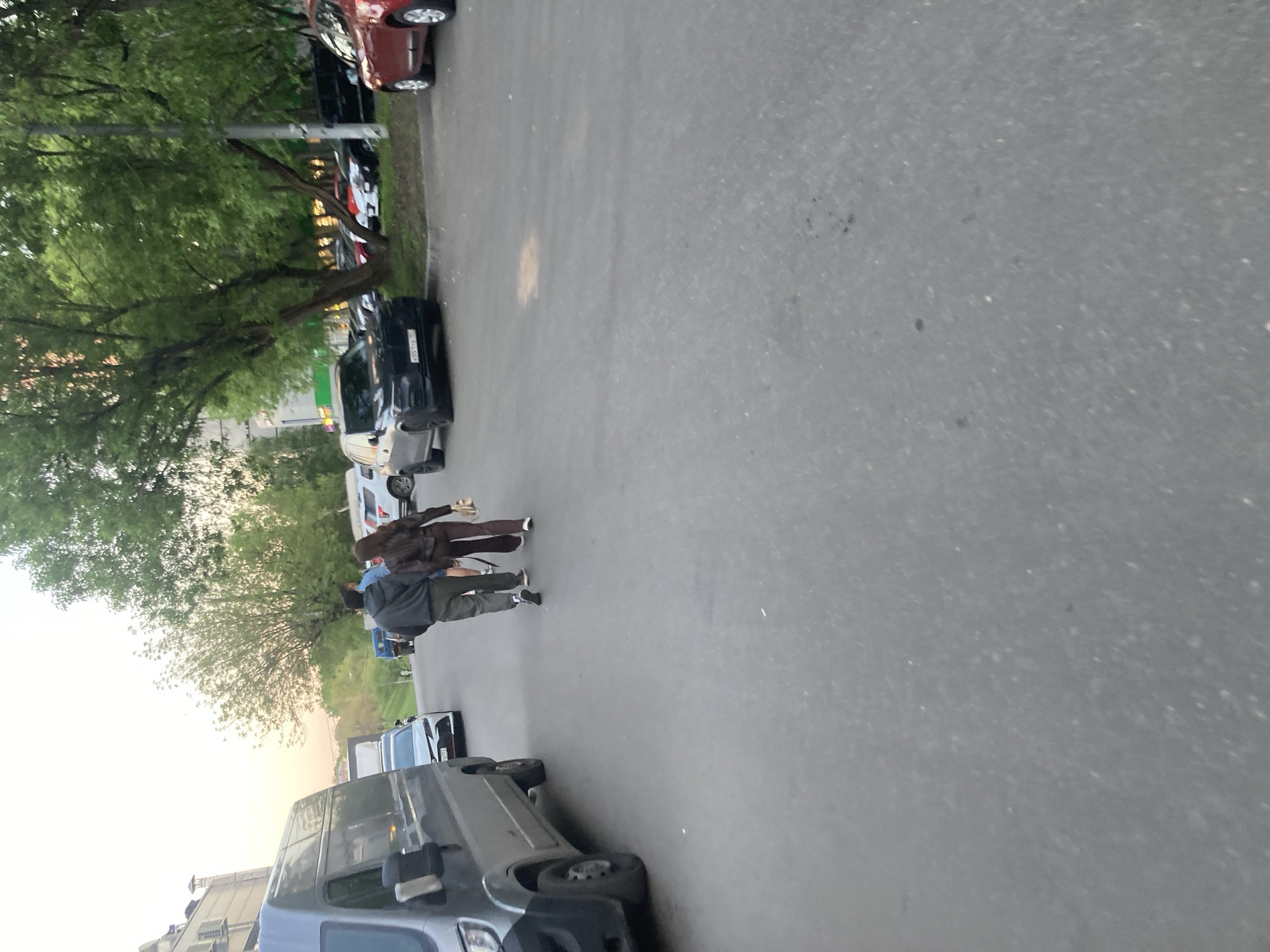 The image shows an outdoor scene in what appears to be a residential area. Two individuals are walking on a paved road alongside parked cars. The individual in the front appears to be wearing a brown jacket, while the individual behind them is in a grey jacket. Trees are visible on the left side of the image. The location appears to be in the city of Moscow, Russia.