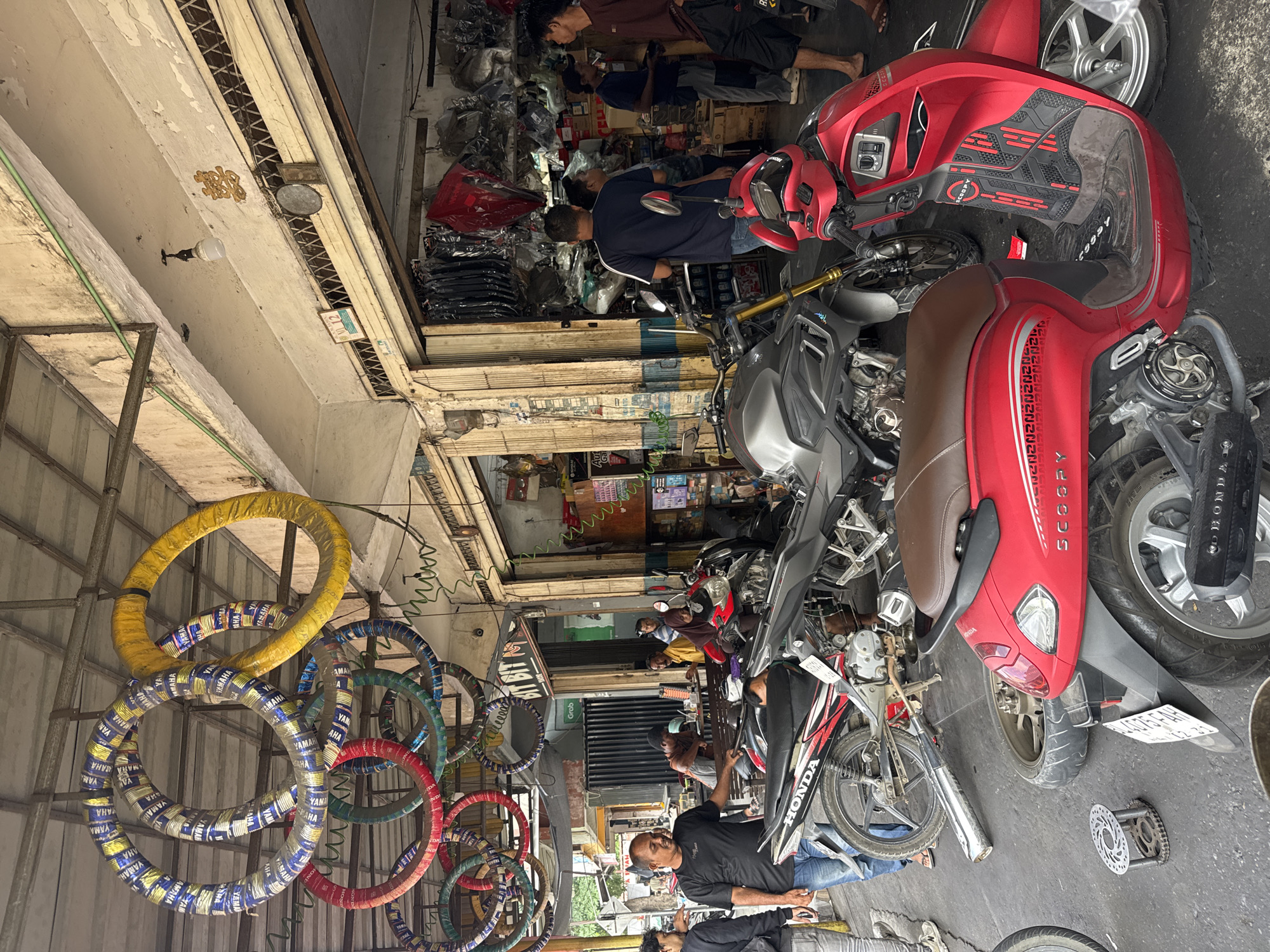 This image depicts a bustling motorcycle repair shop in Banda Aceh, Indonesia. The shop is filled with various motorcycles, including a red Honda Scoopy in the foreground and a grey motorcycle behind it. Tires are prominently displayed, with several large motorcycle tires hanging from the ceiling in colorful packaging.
Several people are present, suggesting a busy workday. A man in a black t-shirt and jeans stands near the front of the shop, appearing to be an employee or owner, engaging with customers. Other individuals are seen in the background, either working on motorcycles, browsing for parts, or waiting for service.
The shop itself appears to be an open-air establishment, with a covered area protecting the workspace from the elements. The walls are somewhat weathered, with visible signs of paint peeling and general wear and tear, typical of many businesses in Southeast Asia. Shelves filled with motorcycle parts and accessories line the back of the shop. The overall atmosphere is one of a functional, active repair and parts service.