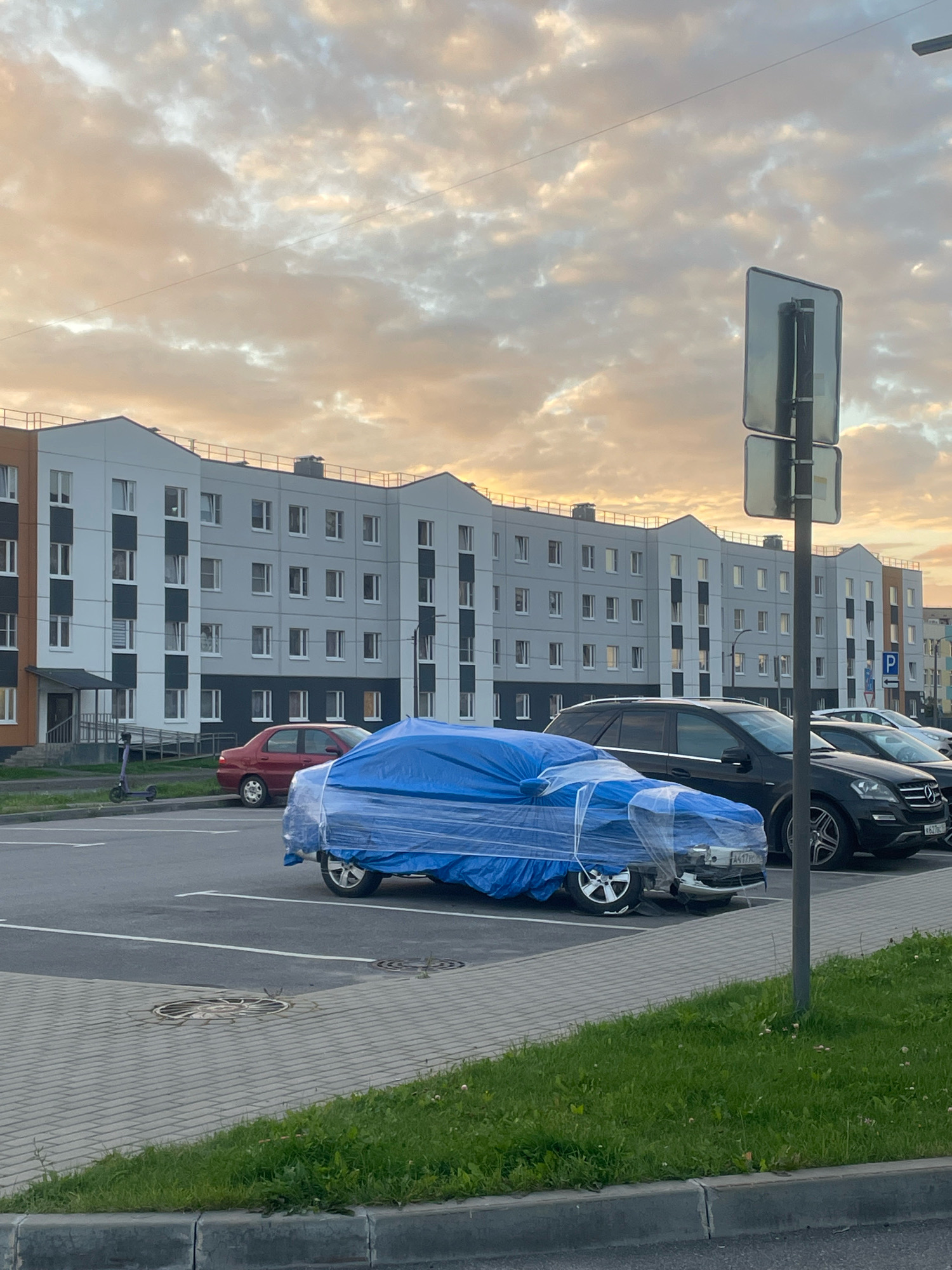 In Gatchina, Russia, on a cloudy day with a soft sunset illuminating the sky, a parking lot is depicted. A multi-story apartment building with a light gray facade and contrasting dark accents stands in the background. In the foreground, several cars are parked. One car, in the center of the image, is covered with a bright blue tarp and plastic wrap. A red sedan is parked to the left of the covered car, and a dark SUV and another car are visible to its right. A scooter is parked near the sidewalk in front of the building. The ground is paved with gray interlocking stones, and a strip of green grass runs along the lower edge of the frame. A traffic sign with two rectangular panels is positioned near the center of the image.