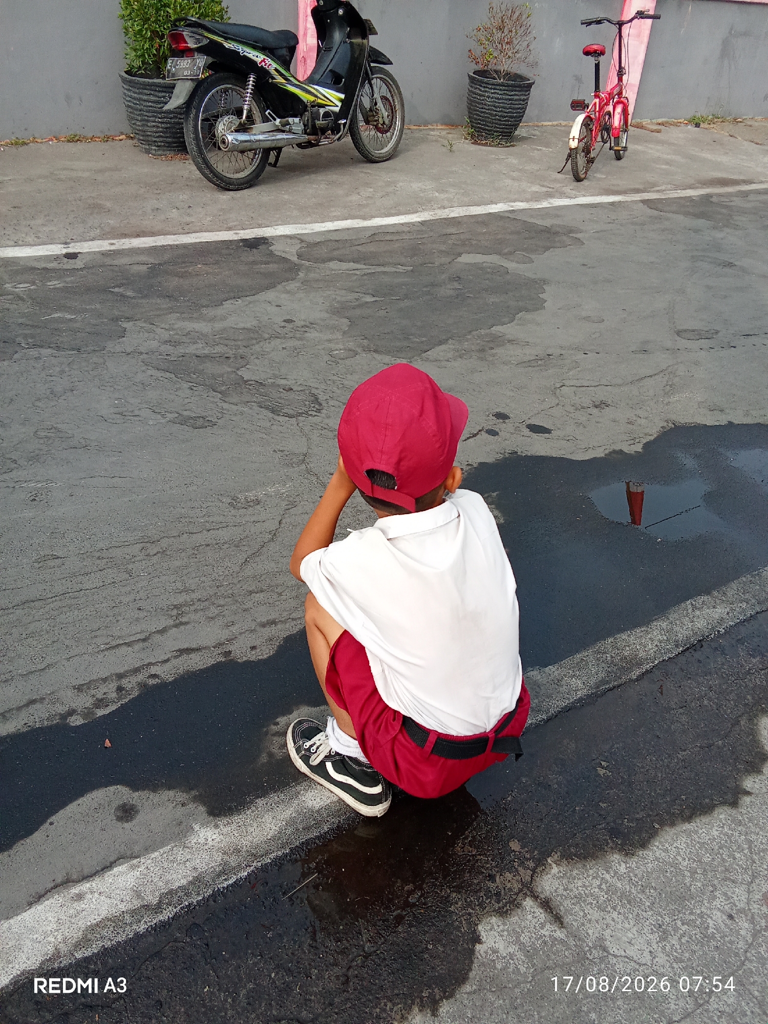 The image depicts a young child, likely a boy, wearing a red cap, white shirt, and red shorts, crouched on wet asphalt. He is looking towards a puddle on the ground, with his hand near his face. In the background, a black motorcycle is parked, and a red bicycle is visible to the right. The setting appears to be an outdoor area, possibly a street or parking lot, with a wall in the background and potted plants. The timestamp "17/08/2026 07:54" is visible in the lower right corner, suggesting the image was taken on August 17, 2026, at 7:54 AM. The location is identified as Cemani, Indonesia.